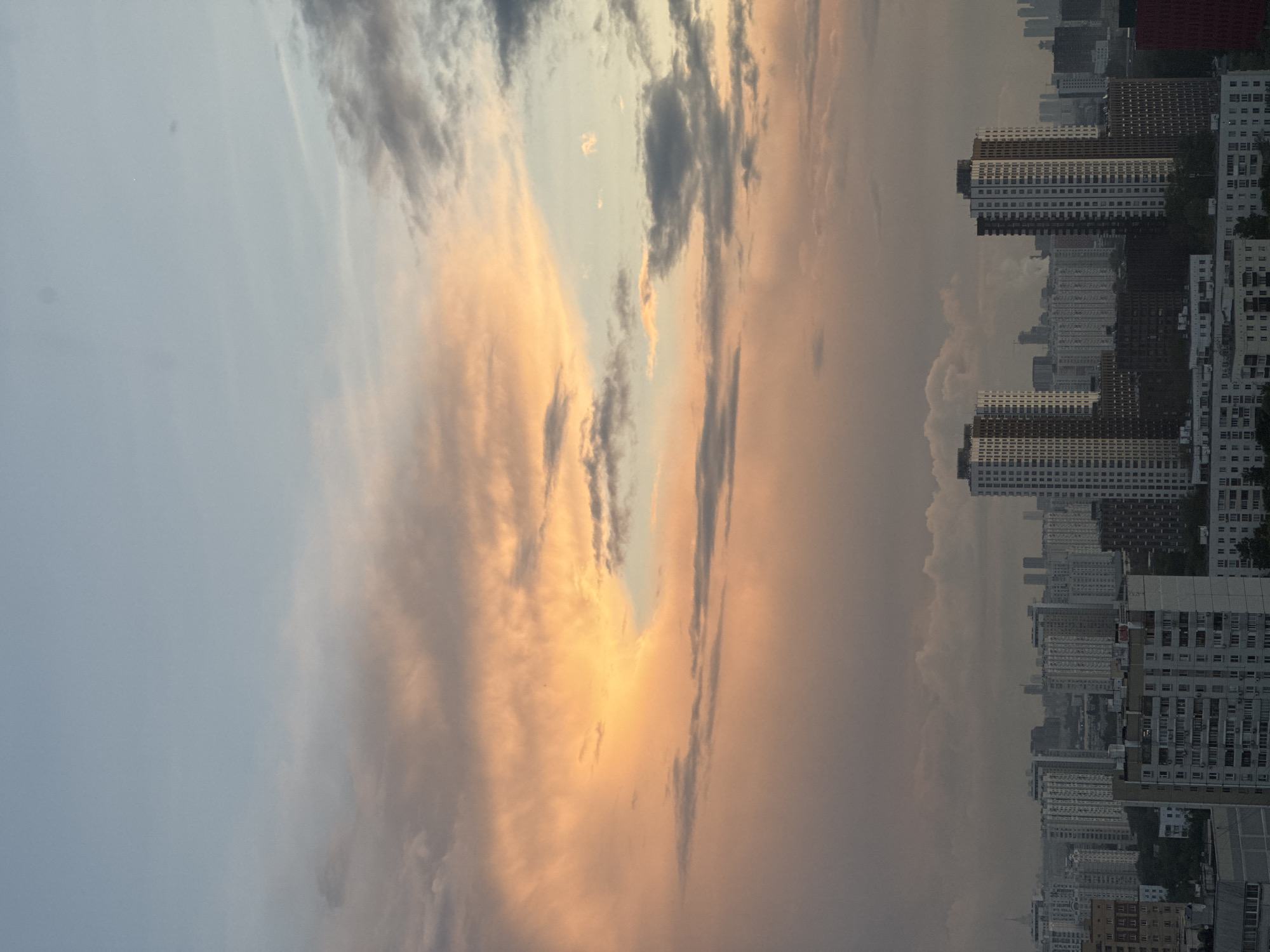 The image depicts a panoramic view of a dense urban cityscape under a dramatic sky. In the foreground, a cluster of multi-story residential buildings dominates the scene, characterized by their repetitive, block-like architecture. These buildings vary in height and color, with some featuring light facades and others darker, more monolithic structures. Some buildings show signs of wear and tear, with visible balconies, windows, and air conditioning units. Lush green trees are interspersed among the buildings, suggesting a developed urban environment with green spaces.
In the midground and background, the city extends further, with a multitude of buildings of varying designs and heights stretching towards the horizon. Some taller, more modern skyscrapers are visible in the distance, interspersed with older, lower-rise structures. Construction cranes are also present, indicating ongoing development within the city.
The sky above is a spectacle of color and texture. Large, billowing clouds are illuminated by a soft, warm light, suggesting either sunrise or sunset. The clouds range in color from pale pinks and oranges to muted grays and blues, creating a dynamic and atmospheric backdrop to the urban landscape. The overall impression is one of a sprawling metropolis bathed in the soft, diffused light of twilight.
While the specific location within Moscow, Russia, cannot be definitively determined from the image alone, the style of architecture, the density of the housing blocks, and the presence of numerous high-rise buildings are characteristic of many residential districts in Moscow. The image captures a common sight in large Russian cities, where vast neighborhoods of apartment buildings are a defining feature of the urban fabric. There are no people or specific actions visible in the image, making it a static, environmental study of the city.