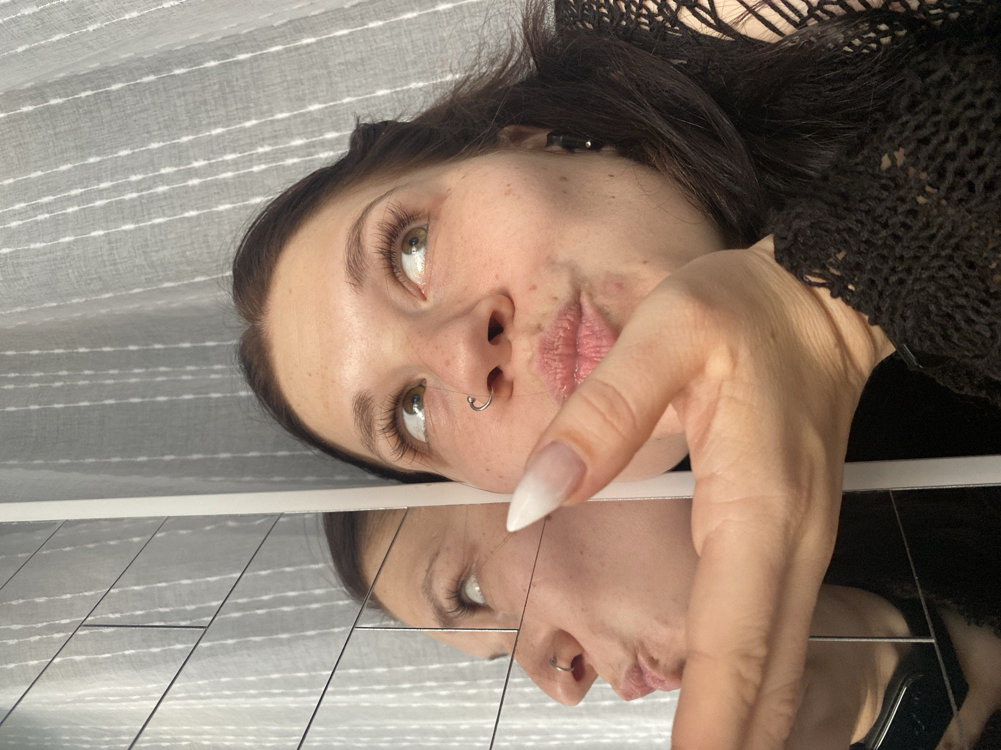 A close-up, high-angle shot captures a young woman with dark hair, light eyes, freckles, and a silver nose ring, reflected in a segmented mirror surface below her. She is positioned horizontally in the frame, looking upwards and to her left, with her lips pursed. Her right hand is raised near her face, featuring a long, pointed fingernail with a white tip fading into a natural base. She wears a dark, possibly crocheted or netted, top.
The background above her is a light-colored, textured fabric, likely a curtain or blind, with subtle horizontal striped patterns. Natural light illuminates her face, highlighting her long eyelashes and the sparkle in her eyes. The reflective surface below shows a clear, inverted image of her face, hand, and the striped background. Visible seams in the reflective surface suggest it is composed of multiple mirror tiles.
The setting appears to be indoors, possibly a domestic environment, characterized by the fabric background and the unique reflective setup. There is no visible text. The image conveys a playful or contemplative mood, captured during daytime in Chaikovsky, Russia.