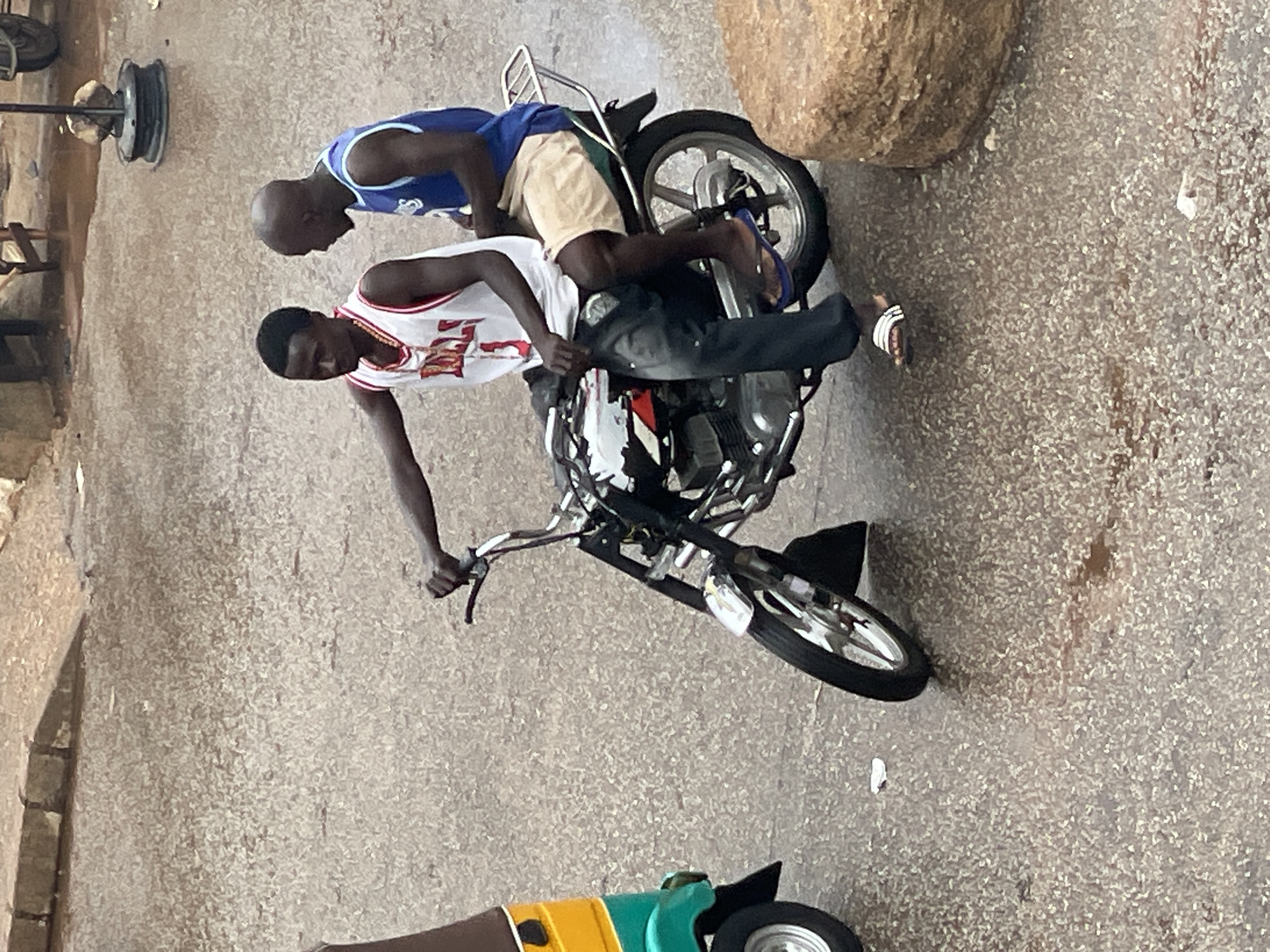 Two young men are riding on a motorcycle on a wet, paved surface in an urban environment. The man in front is driving, wearing a white Bulls basketball jersey and dark pants. The man behind him is wearing a blue basketball jersey and light-colored shorts. They appear to be moving forward. To the left of the motorcycle, a portion of a yellow and green three-wheeled vehicle is visible. The ground is damp, suggesting recent rain. In the background, there are buildings and possibly market stalls, with some debris visible on the ground. A large, round, light-brown object, possibly a rock or a stacked item, is to the right of the motorcycle. The overall impression is a scene of everyday life in a busy city.