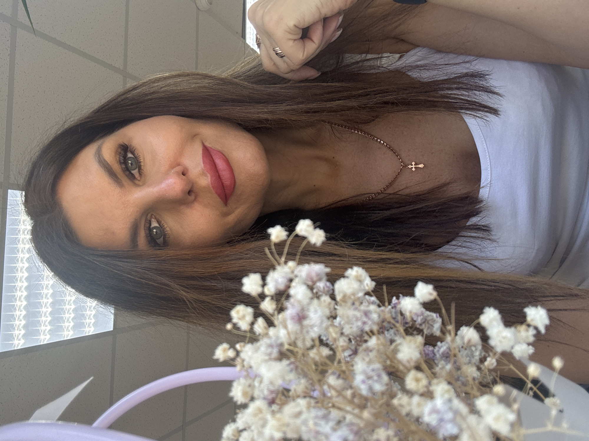 A woman with long brown hair, a fair complexion, and wearing a white top is looking at the camera. She has on red lipstick and is wearing a gold necklace with a cross pendant. The woman is posed next to a small bouquet of white and purple flowers. The background is indistinct but appears to be an interior space with a window on the left. The location is not discernable as being Chaikovsky, Russia, from visual data.