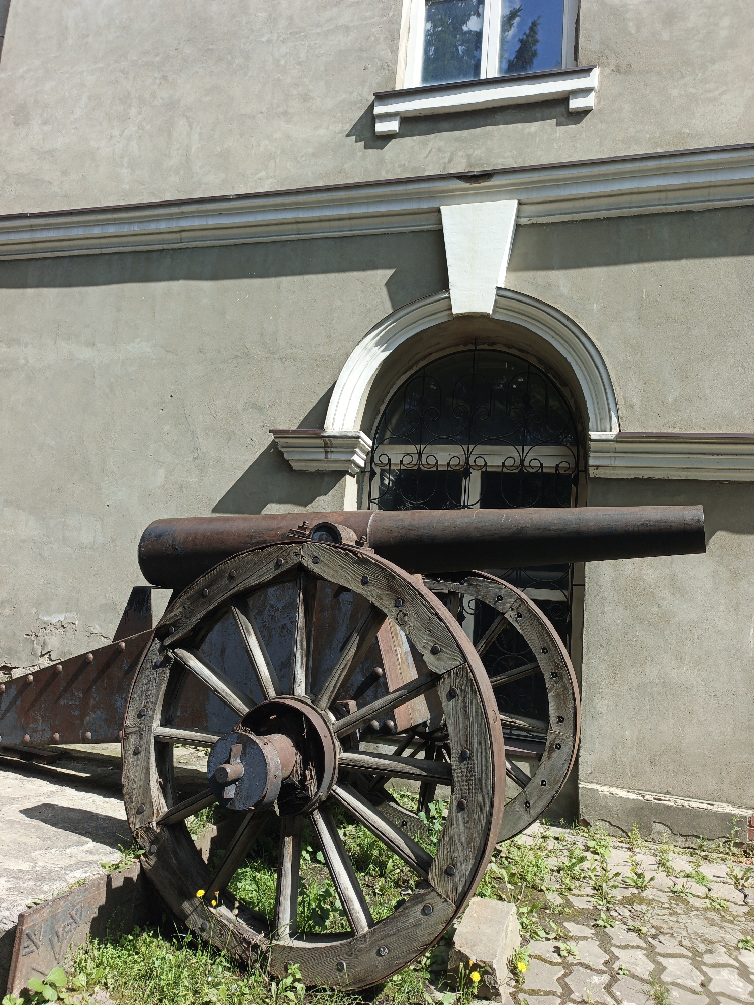 An antique, rusty cannon with large wooden wheels sits outdoors on a paved area with scattered grass, in front of a pale gray building. The building has an arched window with decorative metalwork and a modern white-framed window above it. Decorative white molding runs horizontally across the building's facade. The cannon appears to be a historical exhibit or monument, with no people present. The setting suggests a public space, possibly a museum courtyard or historical site in Zlatoust, Russia.