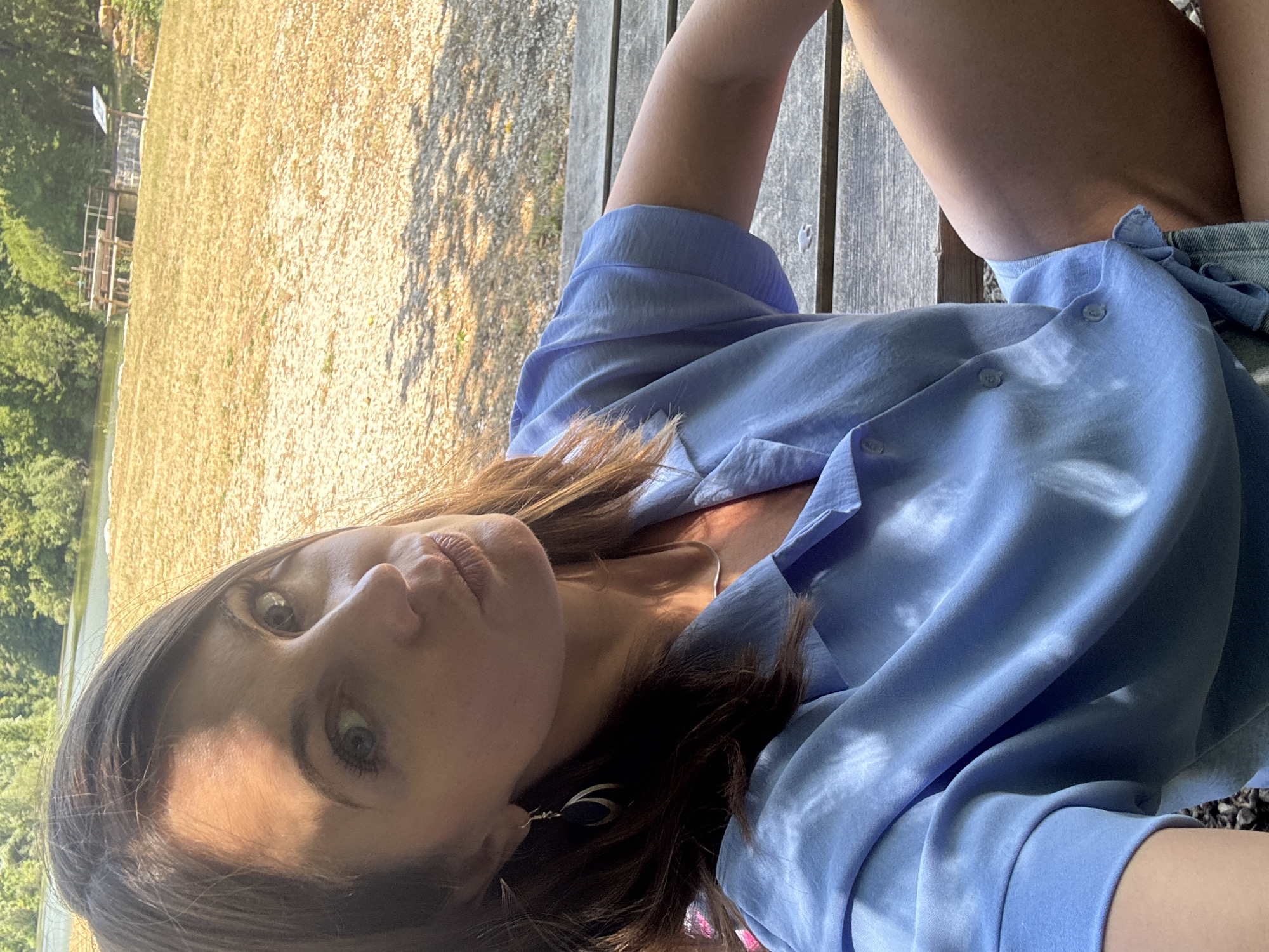 A woman is photographed from the chest up, sitting outdoors on a wooden bench. She has brown hair, blue eyes, and is wearing a light blue button-up shirt and a thin necklace. Her left arm is resting on the bench, and her legs are visible below the frame. The background shows a grassy area, a body of water, and trees. The scene appears to be a park or recreational area.