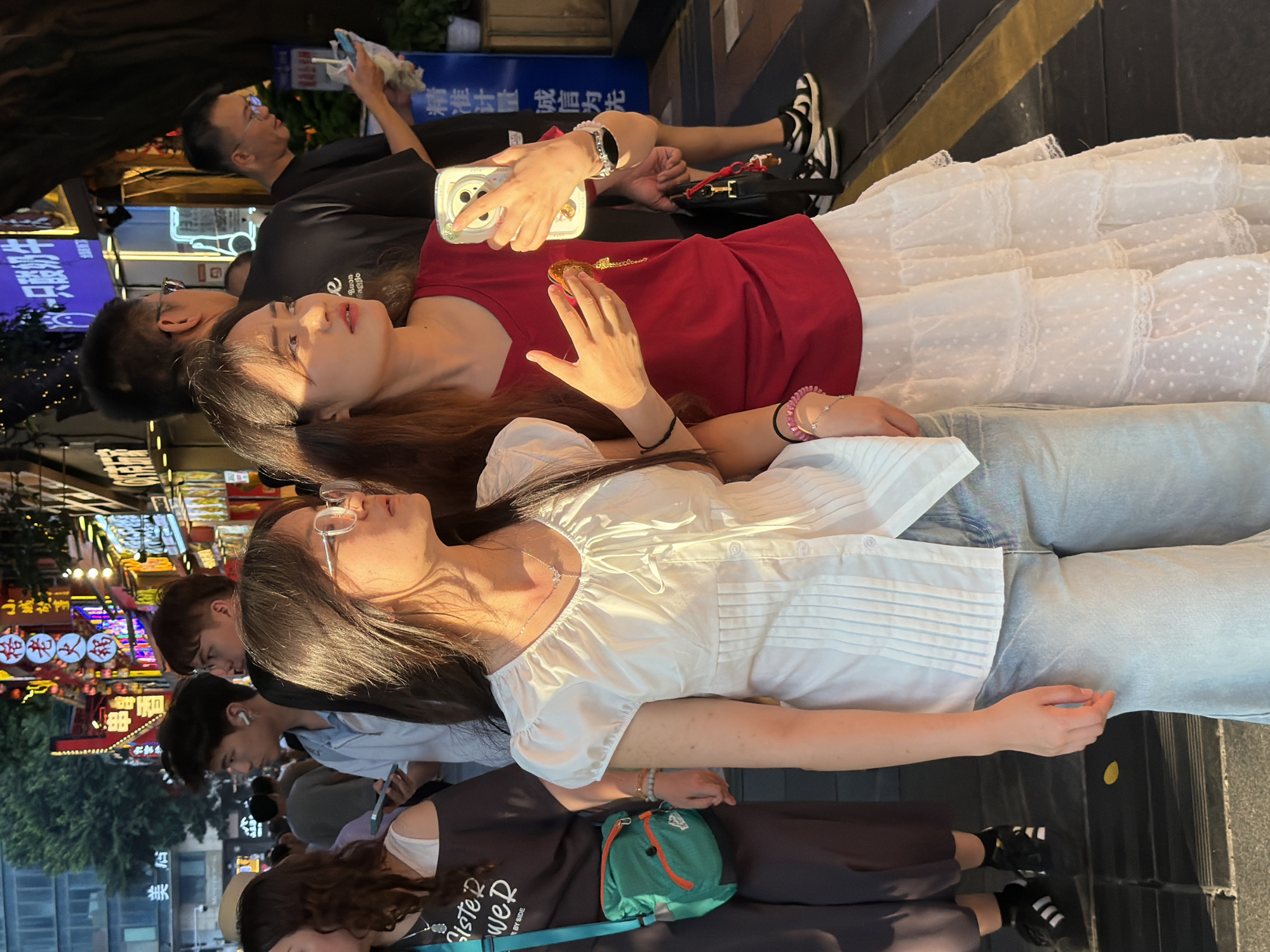 This is a vertical, eye-level, medium shot of several people on a street at what appears to be dusk. The subjects are a mix of males and females, with several women in the foreground. One woman in a red top is holding up a phone to take a picture or video of herself. She is wearing a white, ruffled, polka-dotted dress and bracelets. Beside her, another woman in a white blouse and light blue jeans is looking to her right. The people in the background are mostly out of focus, but one man in a black t-shirt and glasses is visible looking upwards. Neon signs with Chinese characters are visible in the background, indicating a commercial or entertainment district. The text "小城故事" and "精准计量诚信为先" are legible on signs. The scene appears to be a busy pedestrian street, possibly a night market or a street with restaurants and shops. The lighting suggests late afternoon or early evening.
