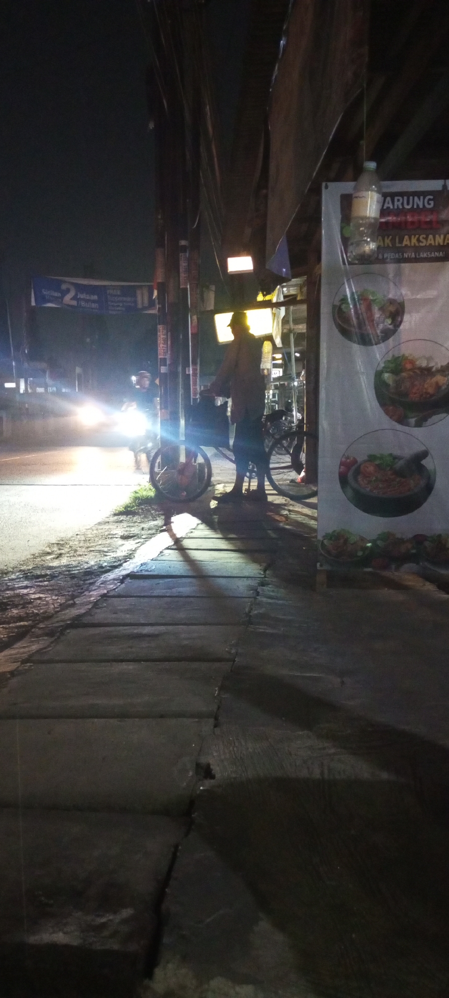 The image depicts a street scene at night in Setu, Indonesia. The foreground shows a concrete sidewalk with visible cracks and unevenness. To the left, a paved road is illuminated by the bright headlights of a passing vehicle, casting reflections on the wet surface. A person is standing on the sidewalk next to a bicycle, holding onto its handlebars. The person is wearing a light-colored long-sleeved shirt, dark pants, and a hat. Their body is silhouetted against the ambient light.
Further back, several utility poles line the side of the road, some with wires strung between them. A banner with Indonesian text and images of food is displayed on the right side of the frame, appearing to advertise a food establishment. There is also a bottle hanging from a string near the banner. The overall lighting is dim, with sources of light coming from street lamps, vehicle headlights, and illuminated signs. The scene suggests a typical urban environment at night, possibly a commercial or residential area.