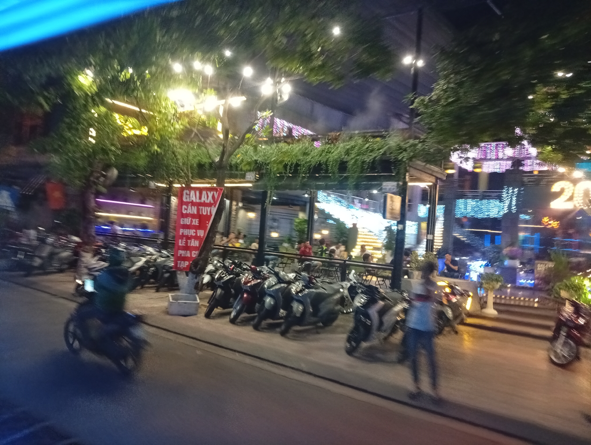 This is a night-time, outdoor scene in Nha Trang, Vietnam, likely a street lined with businesses. In the foreground, a person on a scooter is in motion, with the wheels blurred, indicating movement. The scooter is dark in color. To the left, a red sign with white text is visible, containing the word "GALAXY" prominently. Several parked motorcycles are lined up on the sidewalk.
Further back, the scene opens up to a street with more parked motorcycles and a building with an illuminated facade. Numerous lights, including strings of white and colored lights, decorate the area, suggesting it is a commercial or entertainment district. A few individuals are visible in the background, some seated at tables, while others are standing. The overall impression is of a lively street scene at night in a Vietnamese city. The presence of the word "GALAXY" on the sign and the general atmosphere suggest a commercial area, possibly with restaurants or bars. The blue light visible at the top of the image may be from a street light or a sign.