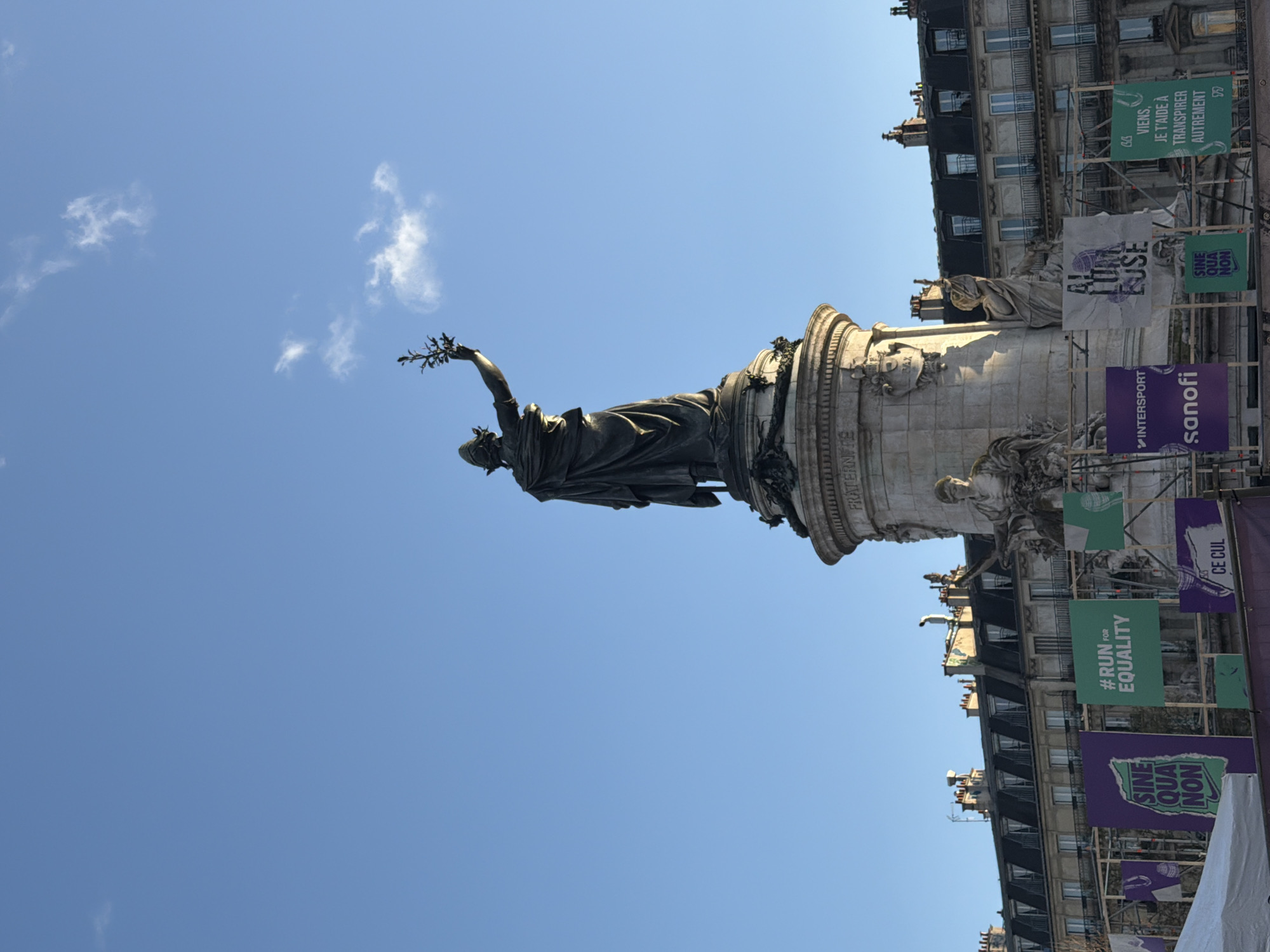The image shows the Place de la République in Paris, France, featuring the monument to the Republic at the center. The statue, depicting a woman holding a branch, stands on a high, circular stone base. Banners with slogans like "#RUN FOR EQUALITY," "SINE QUA NON," and promotional material for "INTERSPORT" and "sanofi" are displayed around the monument. The background includes buildings with traditional Parisian architecture and scaffolding that partially obstructs the view of the structures. The sky is blue with scattered clouds.