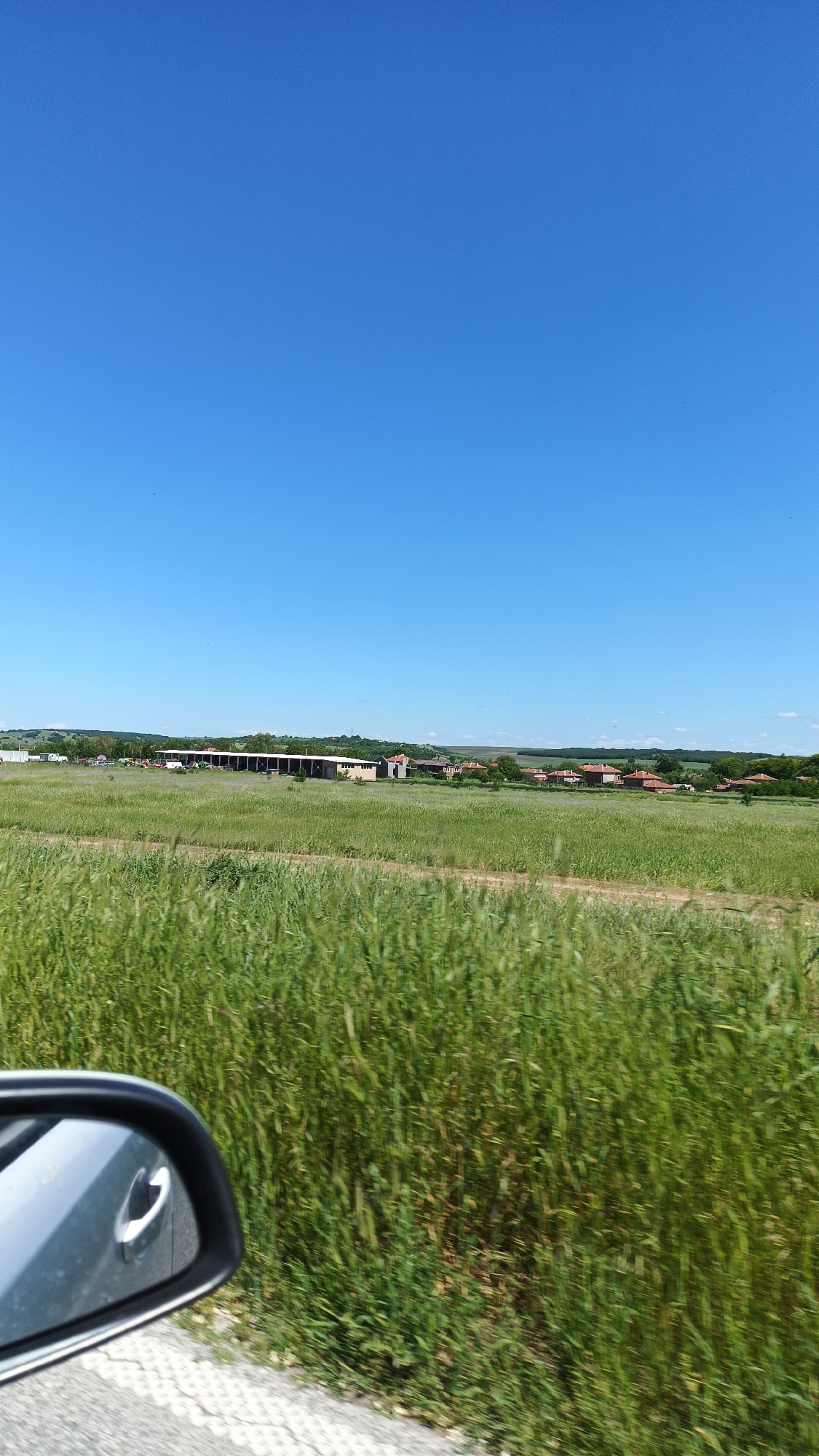 A rural landscape is depicted on a sunny day. A field of green grass occupies the lower portion of the frame, blurred due to motion. In the foreground, part of a side mirror of a car is visible. In the background, there is a village with farm buildings and houses with red roofs. A dirt path separates the foreground field and the village. The sky is clear and blue, taking up most of the upper part of the image. This scene is located in Bogdanitsa, Bulgaria.
