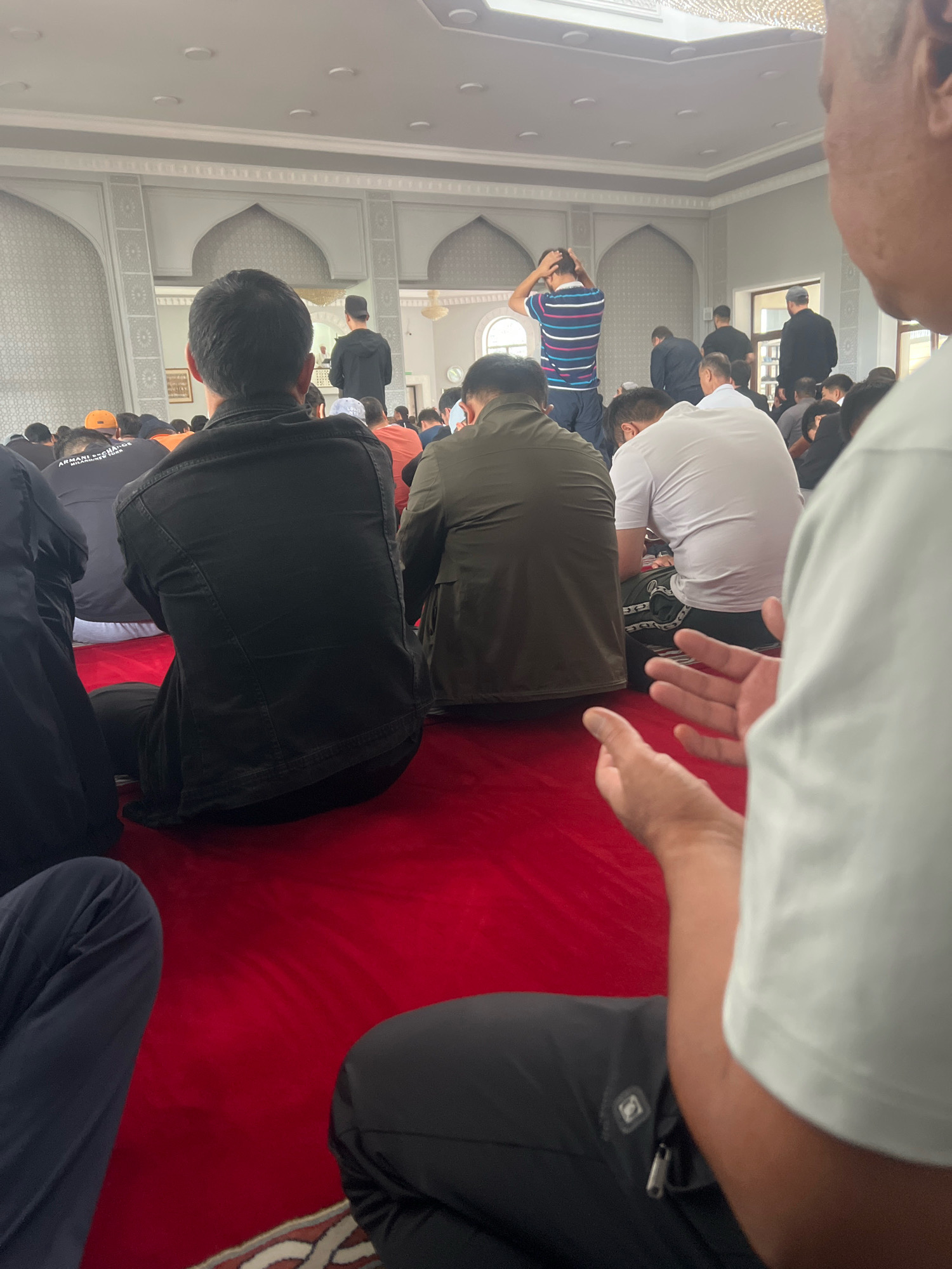 In the city of Almaty, Kazakhstan, a congregation of men are gathered in a mosque for prayer. The mosque features a red carpeted floor, ornate white walls with arched alcoves, and a high, illuminated ceiling. The men are seated or kneeling on the carpet, facing the front of the mosque. Some are in prayer postures, while others are conversing or listening. The attire of the men varies, including traditional Islamic caps, t-shirts, and jackets. The image captures a moment of communal worship in a religious setting.