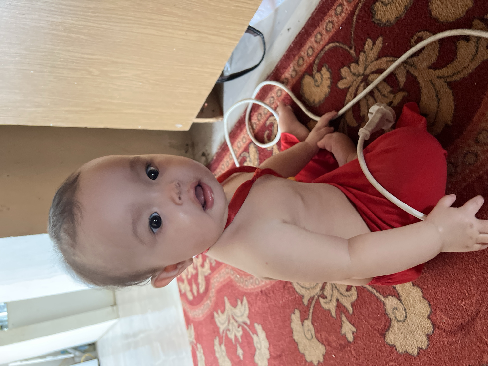 A baby wearing a red garment is lying on a patterned red rug. The baby is facing upwards, with their mouth open and eyes wide. They are positioned next to a wooden surface, possibly a desk or cabinet. A white electrical cord with a plug is visible near the baby's legs and hand, lying on the rug. The scene appears to be indoors, with a carpeted floor and a wooden piece of furniture. The lighting suggests it could be daytime. The baby's expression conveys curiosity or surprise. There is no visible text in the image. The context of Kyzylorda, Kazakhstan is provided.