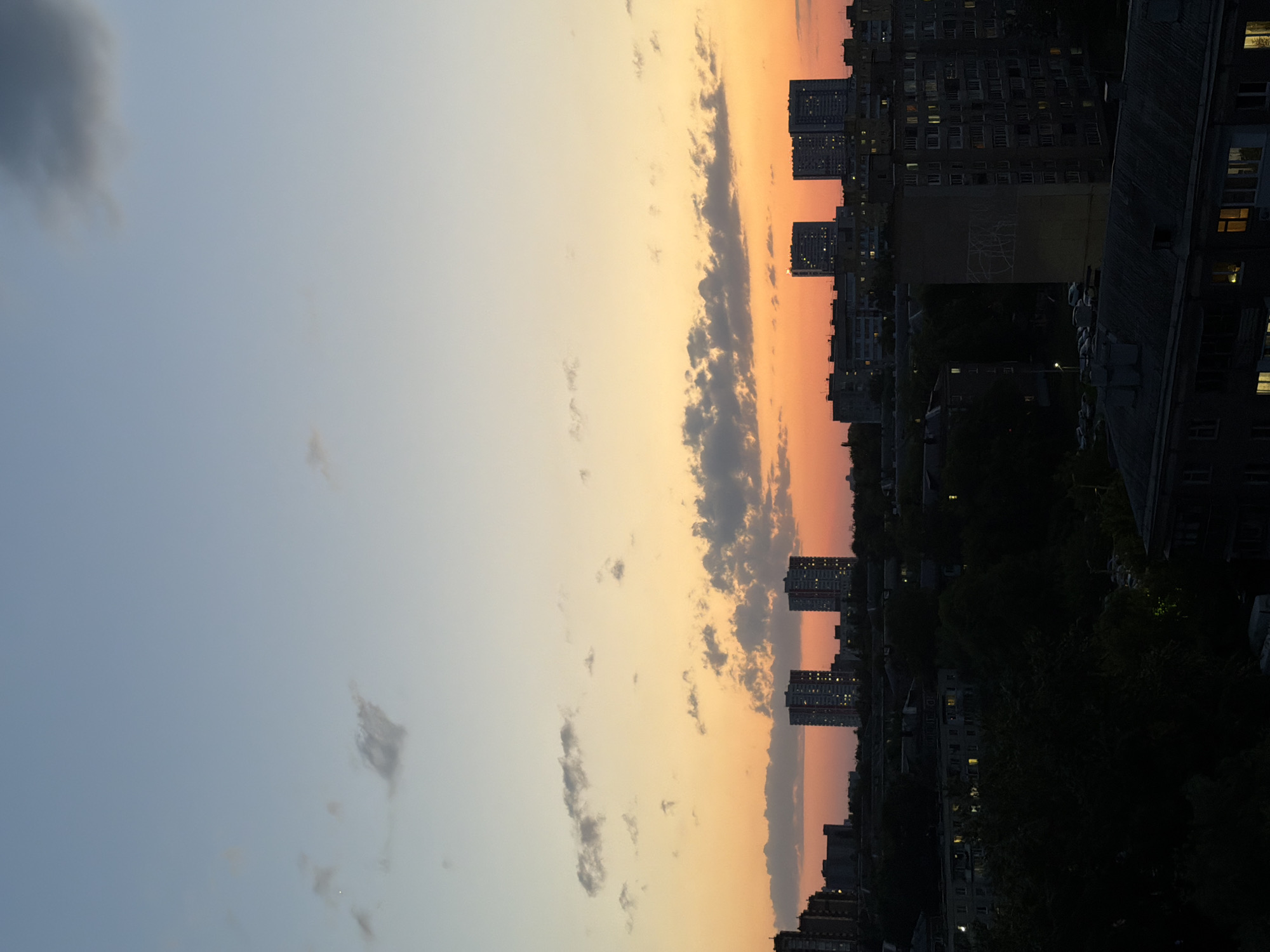 The image shows a cityscape at dusk, with the sky transitioning from blue to orange. Several tall buildings line the horizon, with lights visible in some windows. Clouds are scattered throughout the sky, reflecting the sunset colors. The foreground is darker, with trees and rooftops silhouetted against the fading light.