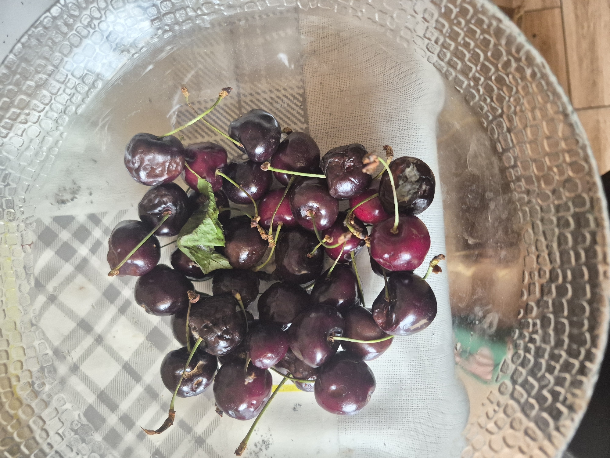 A clear glass bowl filled with dark red cherries sits on a checkered cloth. The bowl has a textured rim. Several cherries are clustered together, with some showing signs of decay. One cherry has a green leaf attached. The background is blurred, but appears to be a wooden surface and a checkered pattern. The image does not provide any information about the city of Yerevan, Armenia.