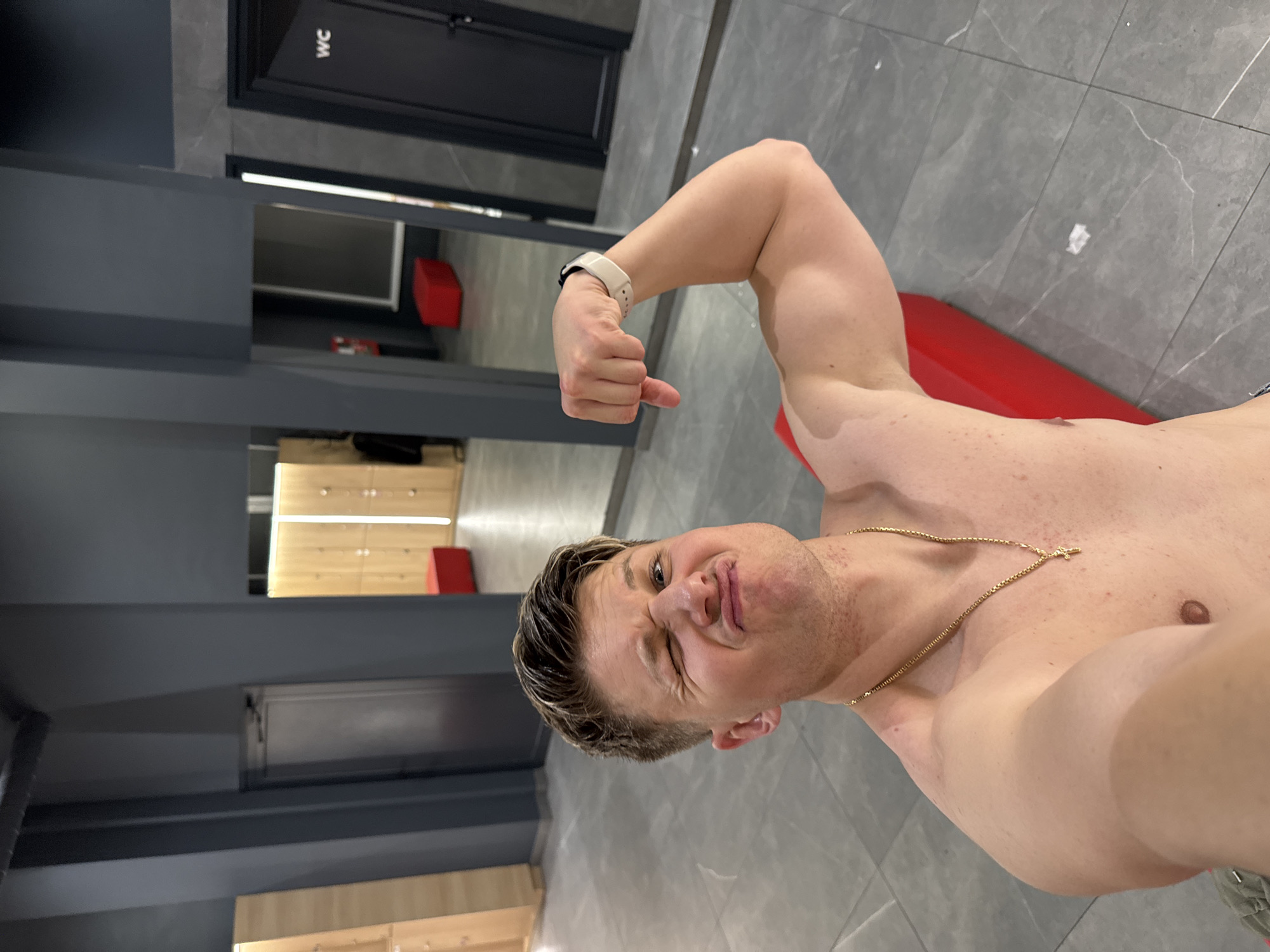 A shirtless man with blond hair and a gold chain is posing for a selfie. He is making a face with one eye squinted and his arm raised, flexing his bicep. He is wearing a white smartwatch. The background appears to be a gym or changing room with gray walls, tile floors, lockers, a red bench, and a door labeled "WC". The location cannot be determined based on the image.