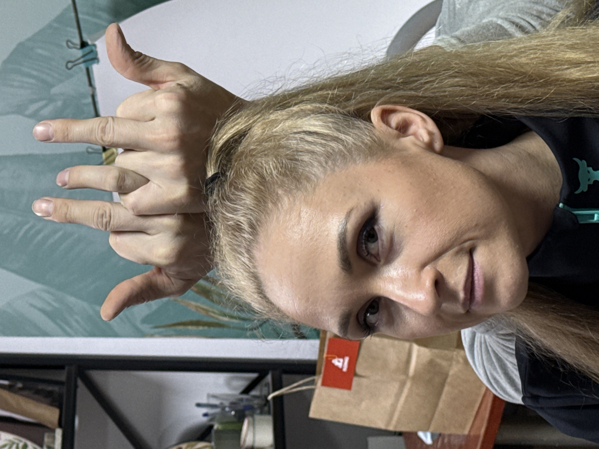 A woman with blonde hair, shaved on the sides, poses at a tilted angle, gesturing with her hands above her head, displaying three fingers on each hand. The background includes a patterned wall, a shelf with various items, and a brown paper bag with a red tag. The location is suggested to be Chaikovsky, Russia, but without further corroborating evidence, this cannot be verified.