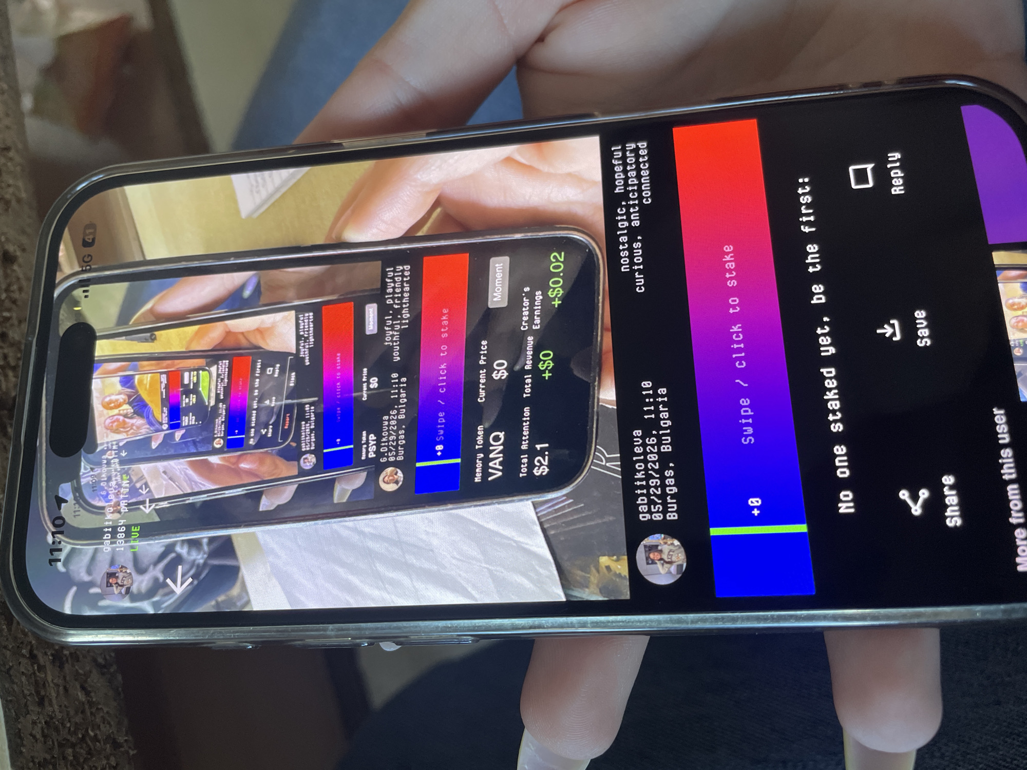 The image shows a person holding a smartphone, displaying a social media interface. The screen depicts a live video feed overlaid with text "gabiikoleva, 05/29/2026, 11:10 Burgas, Bulgaria." There are two tokens shown on the social media interface named PSYP and VANQ with prices $0 and $2.1. The user interfaces include options for staking, sharing, saving, replying, and viewing content from other users. The scene is indoors.