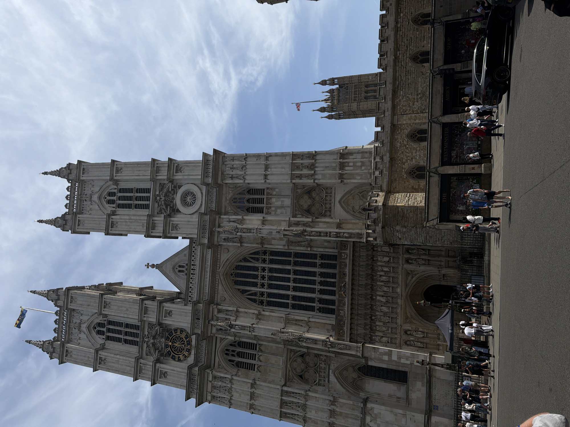 The image showcases a view of Westminster Abbey in London, United Kingdom, angled upwards, with a blue sky and wispy clouds as a backdrop. The abbey's facade is visible in great detail, revealing its architectural features like the pointed arches, stained glass windows, and intricate stone carvings. The clock face and flag can be seen on one of the towers. People are standing near the abbey's entrance, some appearing to be tourists. The abbey's entrance and gift shop, as well as a black car, can be observed. Street paving is also present, indicating an urban environment.
