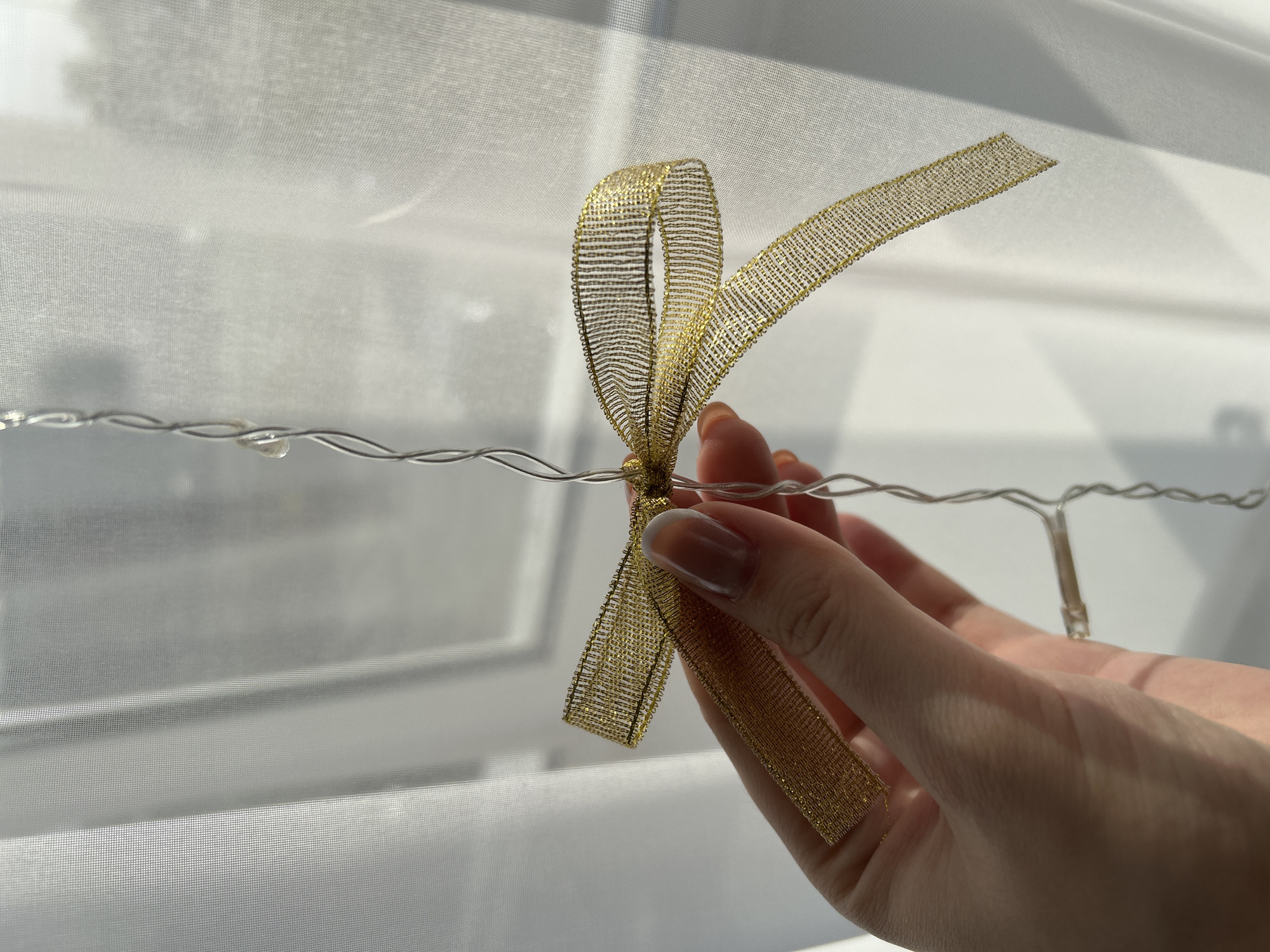 A hand holds a length of string lights, from which hangs a gold ribbon. The background is a white, sheer curtain. The location cannot be determined from the image.