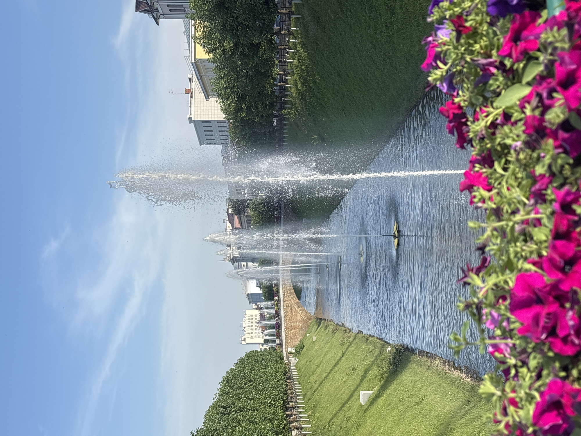 This image shows a canal with several fountains spraying water upwards, located in Kazan, Russia. The canal is flanked by manicured green lawns on both sides, with trees planted along the top of the grassy slopes. In the background, there are buildings and a stone bridge arching over the canal. In the foreground, there are bright pink and purple flowers, slightly out of focus. The sky is blue with a few wispy clouds, suggesting it is a clear day. The scene depicts a park or public space with water features and decorative landscaping.