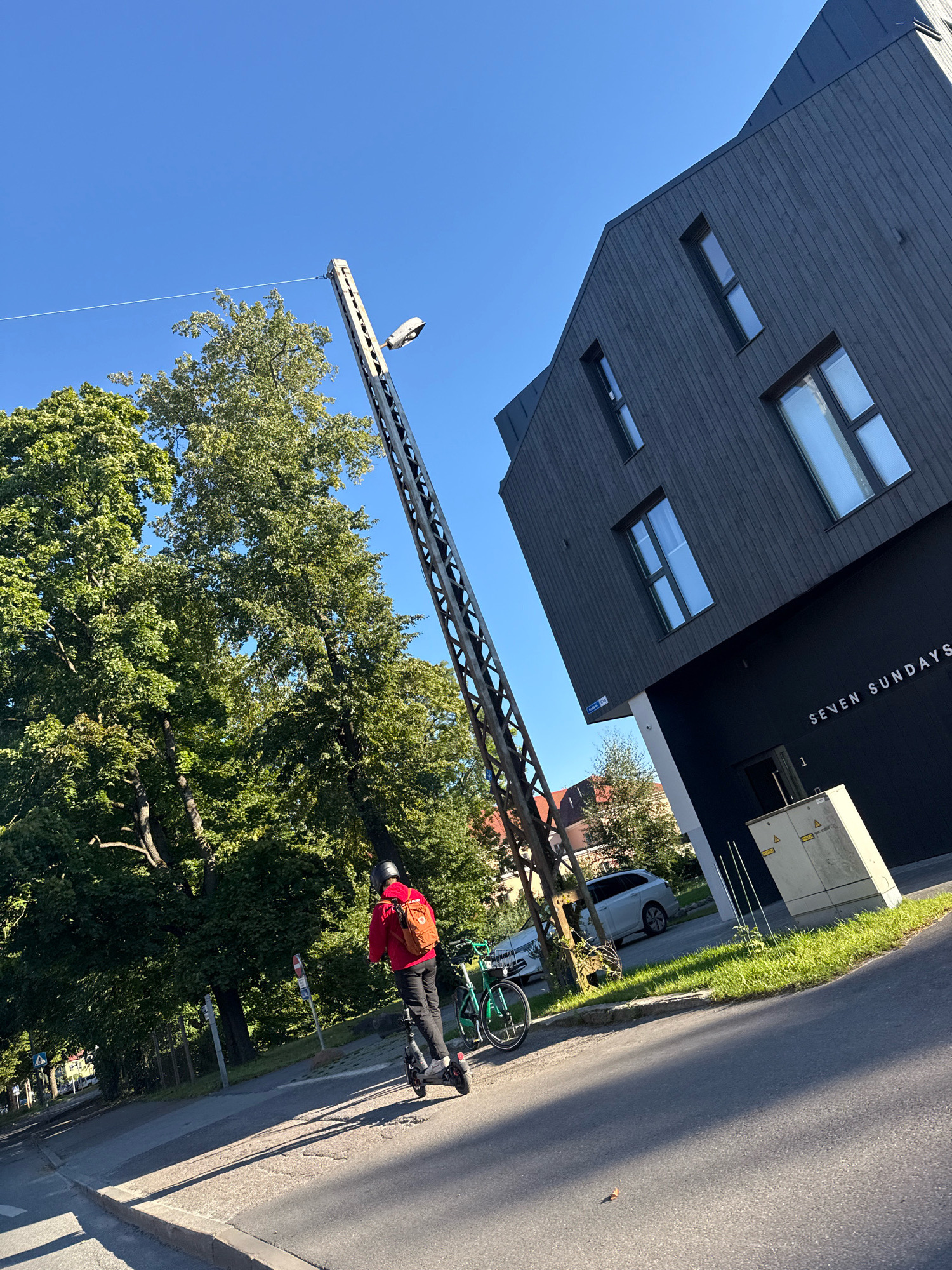 In Tallinn, Estonia, on a sunny day, a person wearing a red hooded jacket and a helmet rides a black electric scooter along a paved road. They have a brown backpack on their back. Beside the road, a green bicycle is parked. To the right of the scene, a modern, dark-colored building with vertical wooden siding and several windows stands under a clear blue sky. The building has the text "SEVEN SUNDAYS" and the number "1" above an entrance. A utility box is situated near the building. To the left of the building and road, a tall metal structure with a light fixture extends upwards, and lush green trees are visible. In the background, a white car is parked on the side of the road.