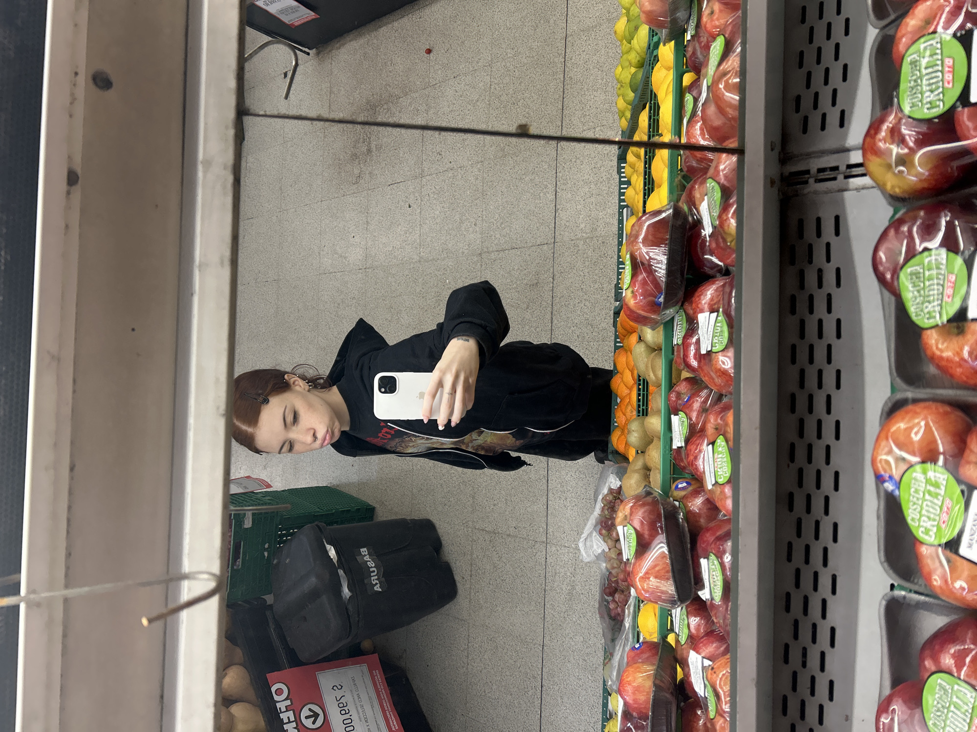 A mirror selfie is displayed in a vertical orientation, showcasing a female individual in a black hoodie, holding a white smartphone, seemingly taking a photograph of herself. The reflection reveals the setting to be a supermarket aisle stocked with produce. The woman is positioned in front of shelves filled with fruits like apples, oranges, lemons and kiwis, packaged in plastic wrap. To her left, behind her, are stacked black bags. Near the bags are two green crates and a sign with red and white text, partially obscured. The environment suggests a typical urban supermarket setting, the name indicates it is located in Buenos Aires, Argentina.