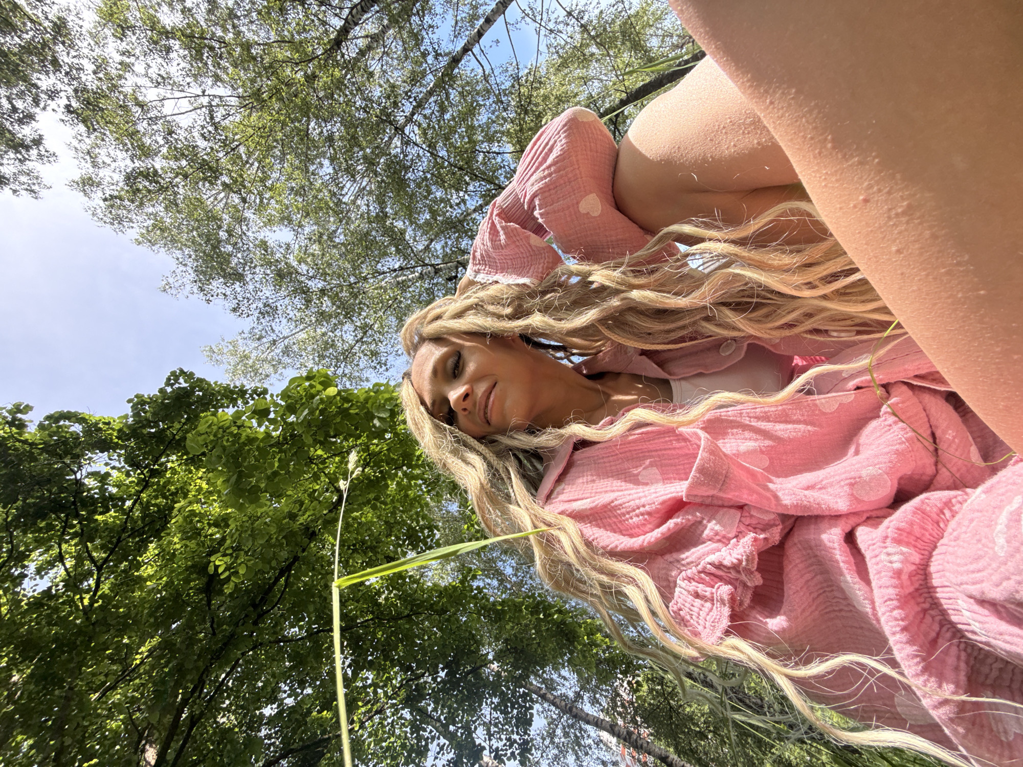 The image captures a woman in a low-angle shot, seated or reclining outdoors in Rusanovo, Russia, under bright daylight. She possesses long, wavy blonde or light brown hair, possibly featuring dreadlocks or similar textured elements, framing her face. She wears a loose-fitting, crinkled pink long-sleeved shirt adorned with small white heart patterns, layered over a white tank top or camisole. Her right arm is bent, with her hand positioned near her head. Her face is turned slightly upward and to the right, displaying a calm expression with partially visible eyes and defined eyelashes. The woman's bare skin on her arms and legs, visible in the foreground, exhibits texture, possibly goosebumps or small epidermal bumps, intensely illuminated by direct sunlight. A single, slender blade of light green grass extends diagonally from the upper left across the frame. The background reveals a clear blue sky interspersed with various trees, including several birch trees with distinctive white bark on the right, and other trees with dense green foliage, suggesting a natural, possibly park-like or wooded, environment.