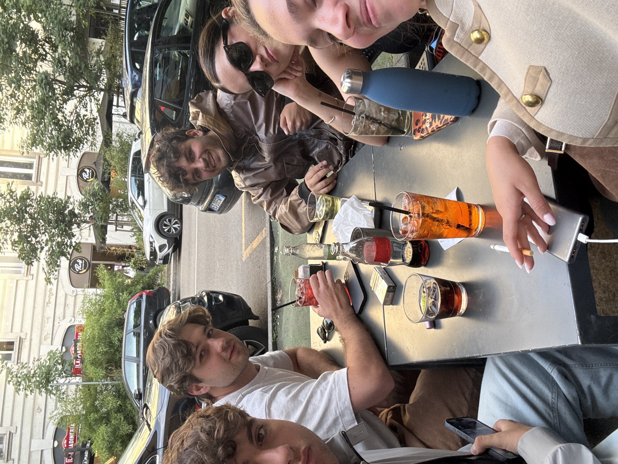 A group of people are seated at a table on an outdoor patio, likely at a bar or cafe. From top to bottom, there are 5 people at the table. A woman in a beige jacket with gold buttons has a phone in one hand and a cigarette in the other. Next to her is an orange cocktail with a straw. To her left is a person in sunglasses, who is next to a man in a brown leather jacket. On the table in front of them are drinks, including a blue water bottle, an unlabeled glass bottle and drinks with straws. In the foreground on the bottom are two men. On the table in front of them are cocktails, a pack of cigarettes, a mobile phone and a pair of sunglasses. The table is made of metal and shows signs of wear and tear. Behind the group, the street shows parked cars and buildings. A sign for the establishment "La Piadineria" is visible in the background. Given the sign and overall visual context, the location is likely in Milan, Italy.