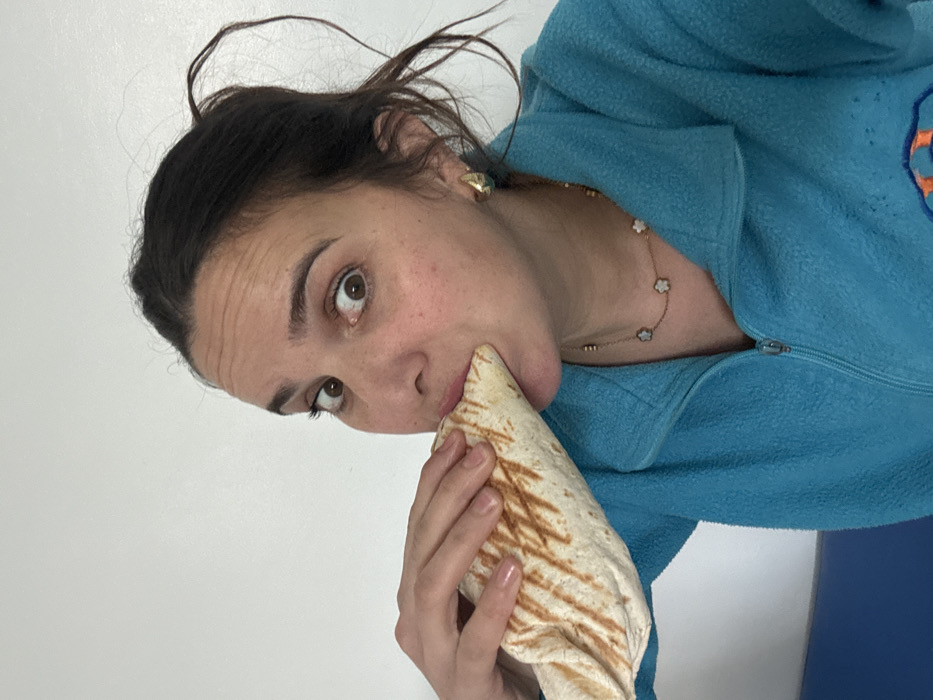 A woman with brown hair pulled back eats a burrito. She wears a light blue fleece jacket over a necklace with flower charms. Her brown eyes look toward the camera. She wears small gold hoop earrings. The burrito has grill marks and is held in her left hand, angled towards her mouth. The background is a plain white wall. A sliver of blue is visible at the bottom right of the image. The photo is rotated counter-clockwise.
