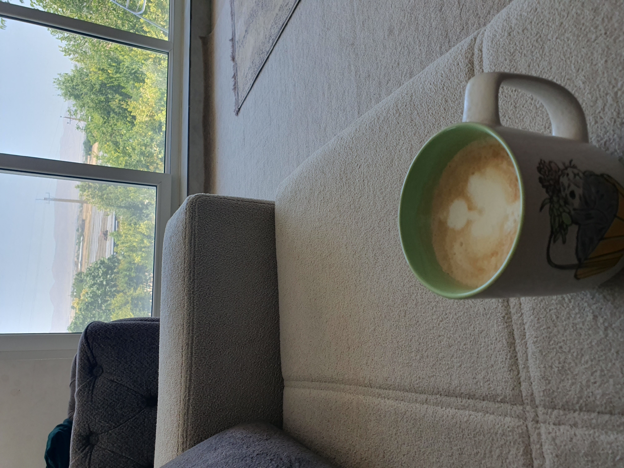 A light-colored textured sofa is in the foreground, with a mug of coffee with latte art on its surface placed on the cushion. The mug has a green rim and a graphic design on its side. In the background, a large window offers a view of trees and distant mountains, suggesting an outdoor setting. The presence of a sofa and window implies an indoor space, likely a living room or lounge. The overall scene is peaceful and relaxed, with the coffee suggesting a moment of leisure. The location of Kheyrabad, Iran, in the context of this image is not visually determinable.