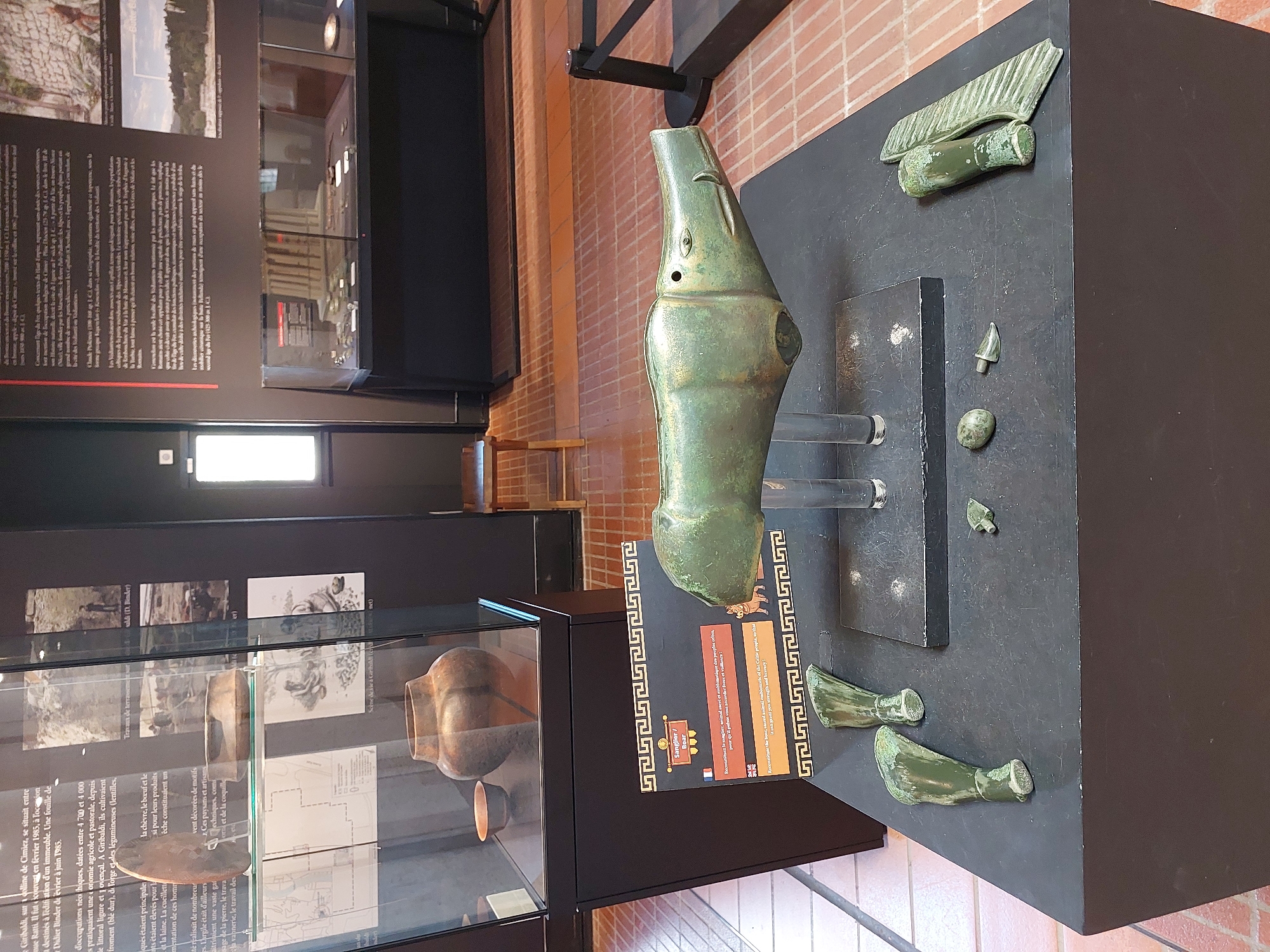 This image depicts an artifact display in a museum, likely the Musée d'Archéologie de Nice-Cimiez, located in Nice, France. The central object is a large, verdigris-patinated bronze sculpture of a boar's head, appearing to be an ancient artifact. It is presented on a dark display platform, supported by clear cylindrical stands. Scattered around the boar sculpture on the platform are several smaller bronze artifacts, including what appear to be animal legs or hooves, a small egg-shaped object, and a pointed piece. To the left of the platform, a black-and-gold placard with Greek-key-style borders provides information about the boar sculpture, with text in French and English. Behind the display, a glass-enclosed exhibit case contains pottery and other artifacts. In the background, museum displays and brick flooring are visible. The lighting suggests an indoor museum environment. There are no people visible in the image.