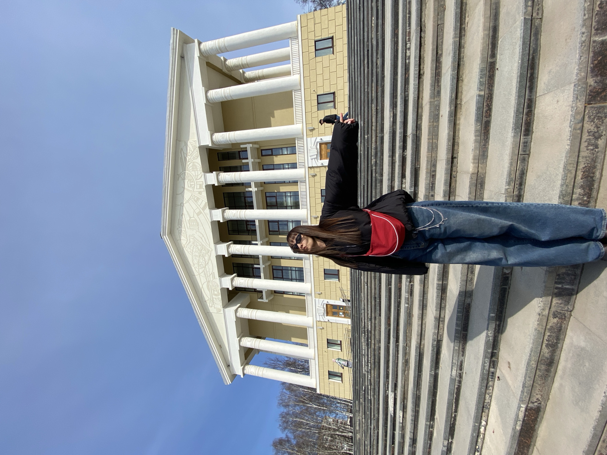 A young woman stands on a wide set of concrete stairs in front of the Votkinsk Palace of Culture in Votkinsk, Russia. She is wearing a black jacket, blue jeans, and a red shoulder bag. She is also wearing sunglasses. Her hair is brown and reaches her shoulders. The woman has both arms raised above her head. The Votkinsk Palace of Culture is a yellow building with white columns. The sky is blue, and the sun is shining. Some bare trees are visible at the bottom of the image.