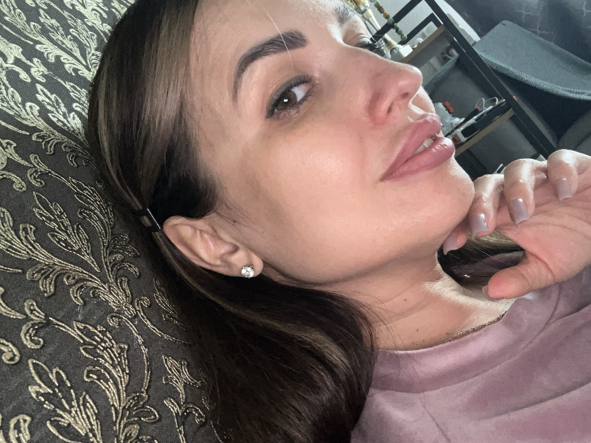 A woman with brown hair and brown eyes is pictured in a close-up shot. She is wearing a light pink velour shirt and a diamond stud earring in her left ear. Her right hand is gently resting on her chin, and her fingernails are painted a light purple color. The background appears to be a couch with an ornate, dark pattern. The lighting is soft and natural.