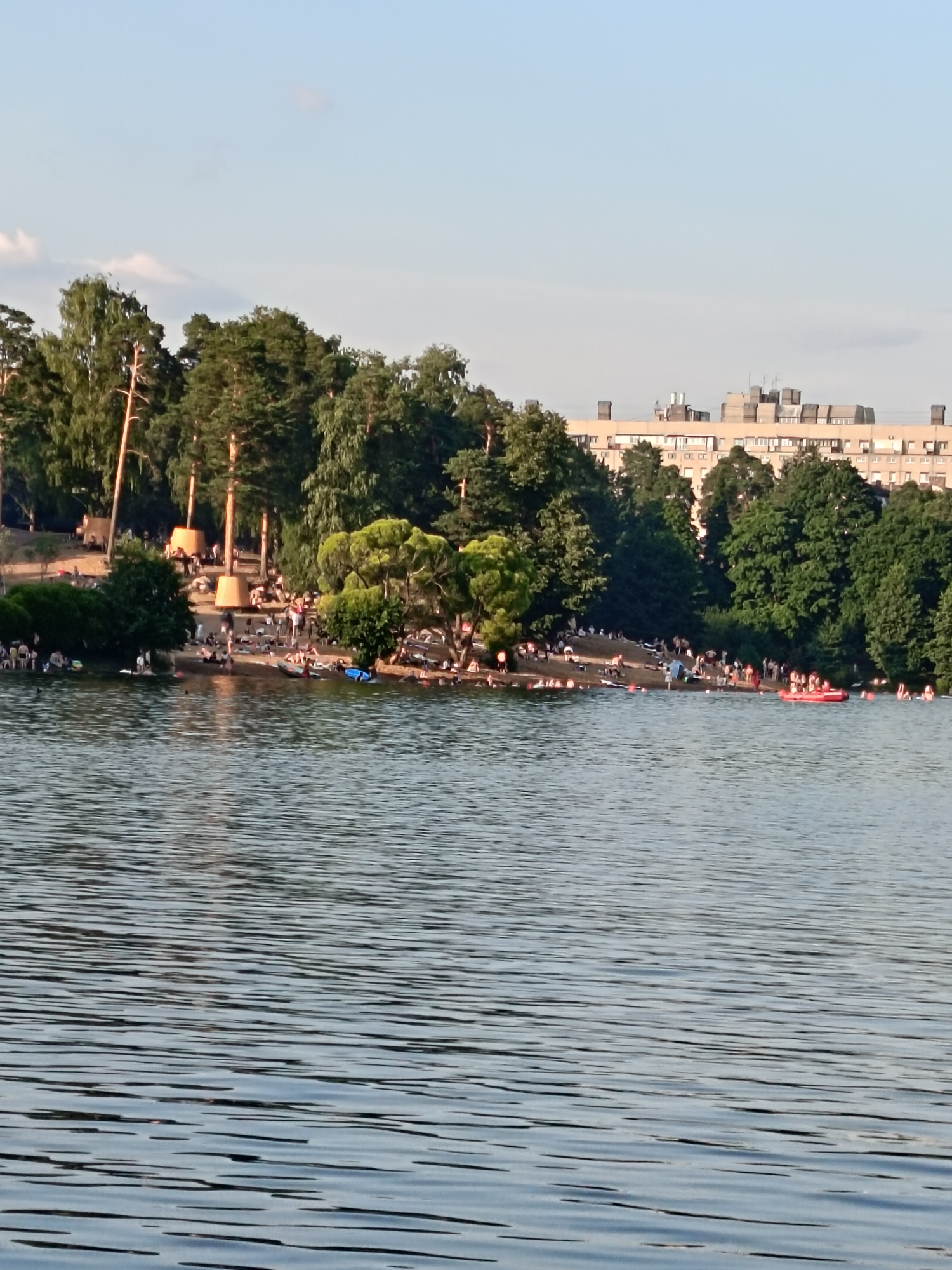 This image shows a wide shot of a lake with a sandy beach on the far side, located in Saint Petersburg, Russia. The beach is populated with many people sunbathing and swimming. There are trees lining the back of the beach, with a large, multi-story building visible in the distance behind the trees. The lake itself is calm with ripples on the surface. In the water, there are several people swimming and a bright red inflatable boat with several occupants. The sky is clear and blue with a few faint clouds.
