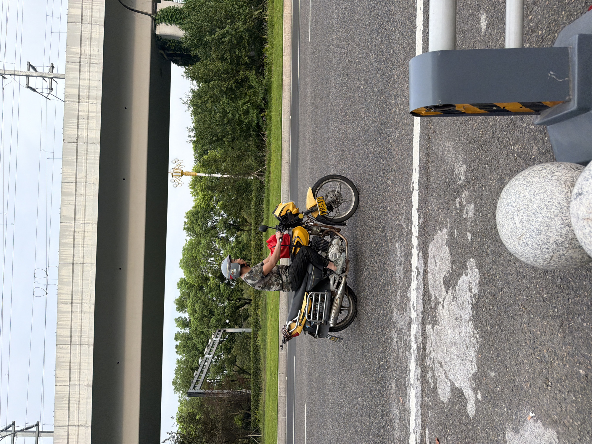 A man wearing a camouflage t-shirt and helmet rides a yellow motorcycle on a paved road, with trees and an elevated road structure in the background. The motorcycle has a red bag attached to the rear rack and a license plate reading "WR538". The road has white lane markings and some wet patches on the surface. In the foreground, a grey bollard with yellow and black stripes is visible, along with two spherical granite objects. The image was taken in the city of Jiangjin, China, under an overcast sky.