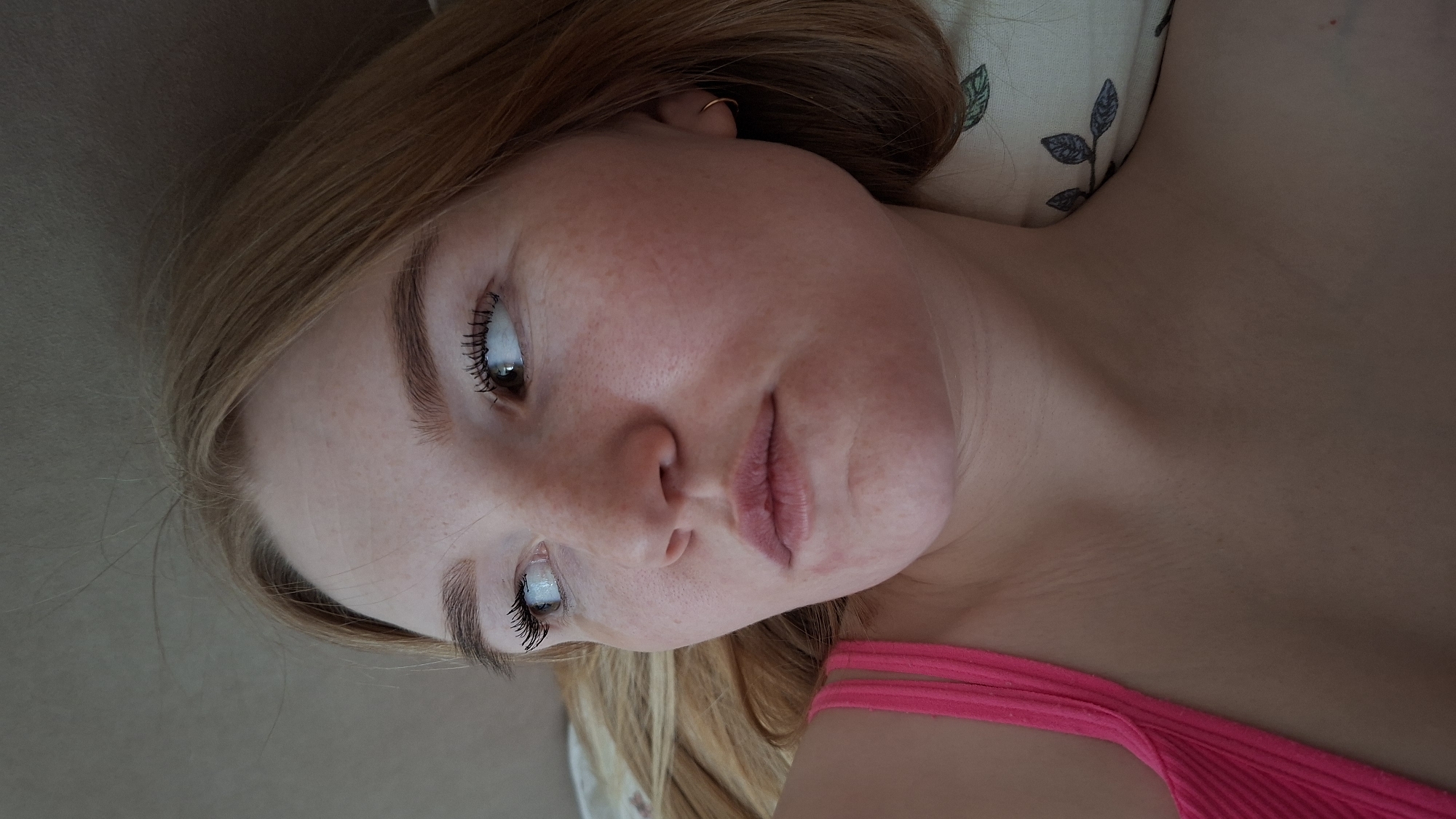 A young, light-skinned woman with freckles lies on her back, her head resting on a pillow with a leaf pattern. She has light brown hair and is wearing an earring in her left ear. She is also wearing a pink top. The location is not discernable from the image.