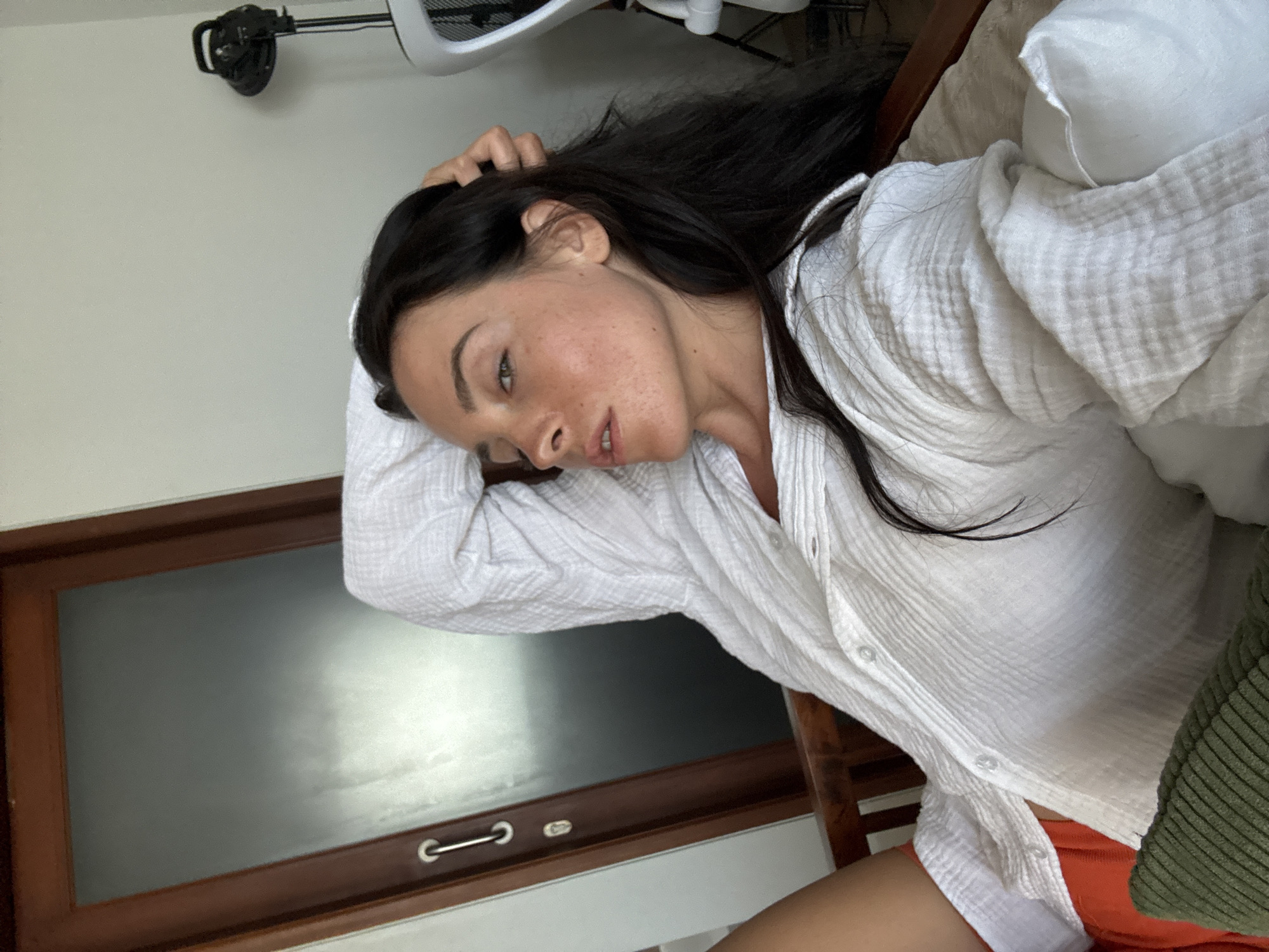 A woman with dark hair and fair skin, wearing a white collared shirt and orange shorts, is lying on a chair with her right hand behind her head. She is looking to her left. The background includes a wooden door with a frosted glass panel, a light-colored wall, and a black fan on a stand. A green pillow is visible in the foreground. The scene appears to be indoors in North Kuta, Indonesia.