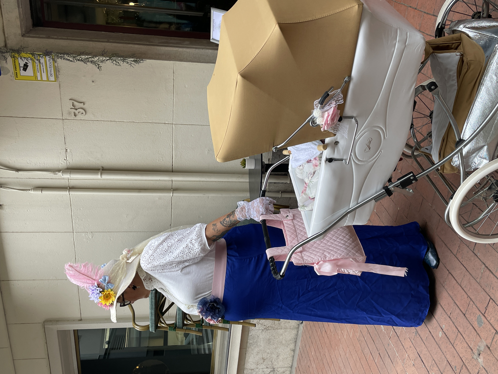 A woman in elaborate period clothing stands next to an ornate baby carriage on a brick walkway. The woman wears a wide-brimmed hat adorned with feathers and flowers, a white lace top, a long blue skirt, and lace gloves. The baby carriage has a white body, a tan umbrella, and silver metal wheels. The number "37" is visible on the building behind the woman, along with a surveillance camera sign. The scene is likely set in Terrassa, Spain.
