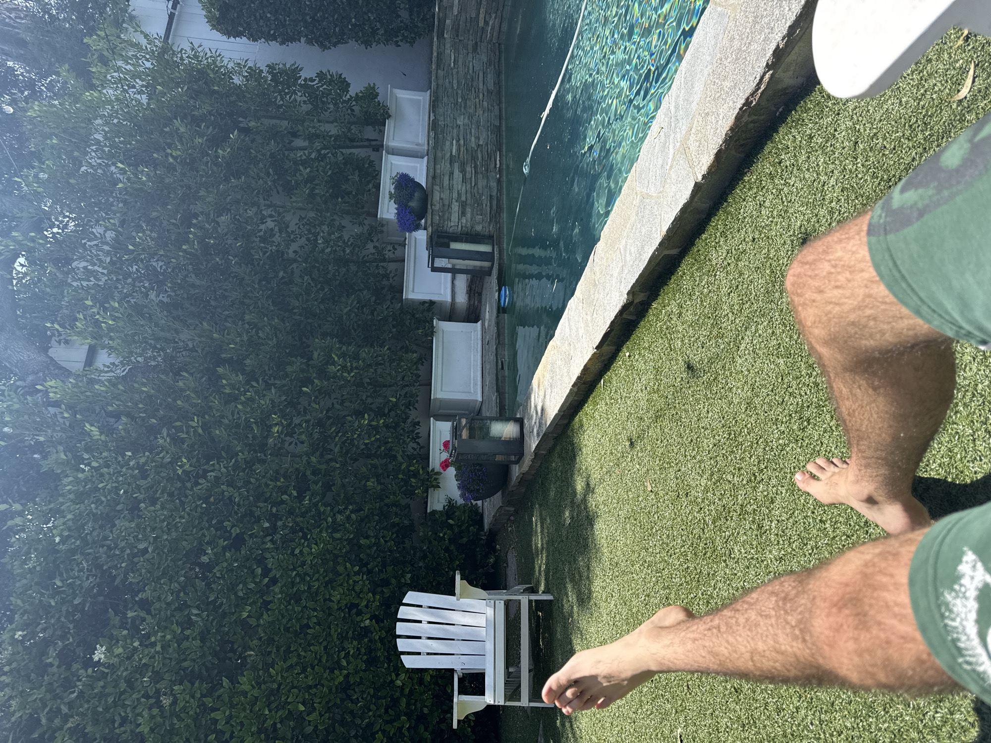 A daytime, first-person perspective image depicts a person sitting in an outdoor space, presumably in the city of Los Angeles, United States of America. The person's legs are visible, clad in green shorts, with bare feet resting on a green, artificial turf. To the left, a dense green hedge lines the edge of the property, with tiered white planters intermittently placed. A white Adirondack chair is positioned near the hedge. Adjacent to the turf is a stone-lined swimming pool with clear, turquoise water reflecting sunlight. A pool cleaning device floats in the pool. The angle suggests the image was taken while seated, looking towards the pool and the garden area.