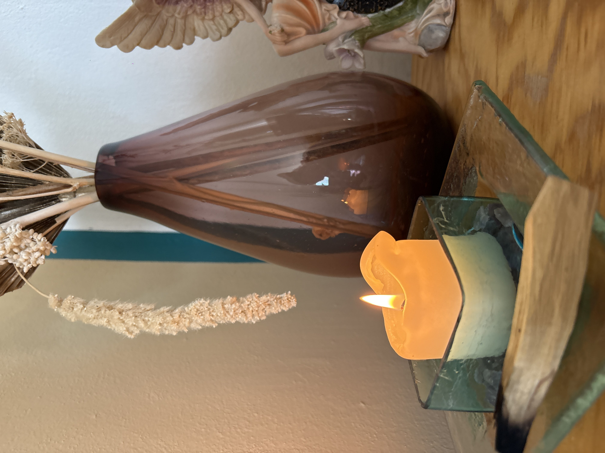 The image displays an indoor still life arrangement, rotated 90 degrees clockwise from its upright position. A prominent, tall, brownish-purple glass vase holds an arrangement of dried botanical elements, including long, thin stems and a fluffy, light-colored seed head extending horizontally.
To the right of the vase, a lit, light yellow candle with a visible flame is nestled within a geometric glass candle holder, which appears to be constructed from several angled pieces of translucent green glass. In the upper background, partially visible, is a decorative figurine, possibly depicting an angel or a classical figure, with visible wings and draped fabric.
The setting includes a light-colored wall, a horizontal teal stripe, and a wooden surface that the objects are resting on. The warm glow of the candle adds to a peaceful and decorative ambiance. Reflections are visible on the glossy surface of the vase. There is no visible text in the image.