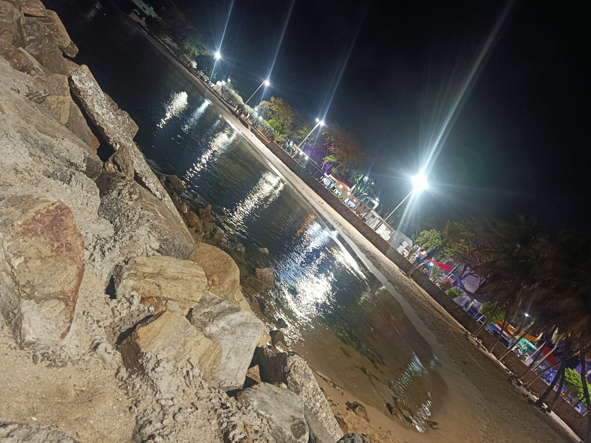 The image depicts a night scene at Juan Griego, Venezuela. A large cluster of rocks sits on the left, descending towards a beach and water. Streetlights illuminate the shore, reflecting in the water. The lights line a beach boardwalk, trees and possibly buildings are visible in the distance. In the far right, palm trees with colorful decorations are visible. There are no people visible. The scene is dark with the exception of the illuminated areas by the streetlights.