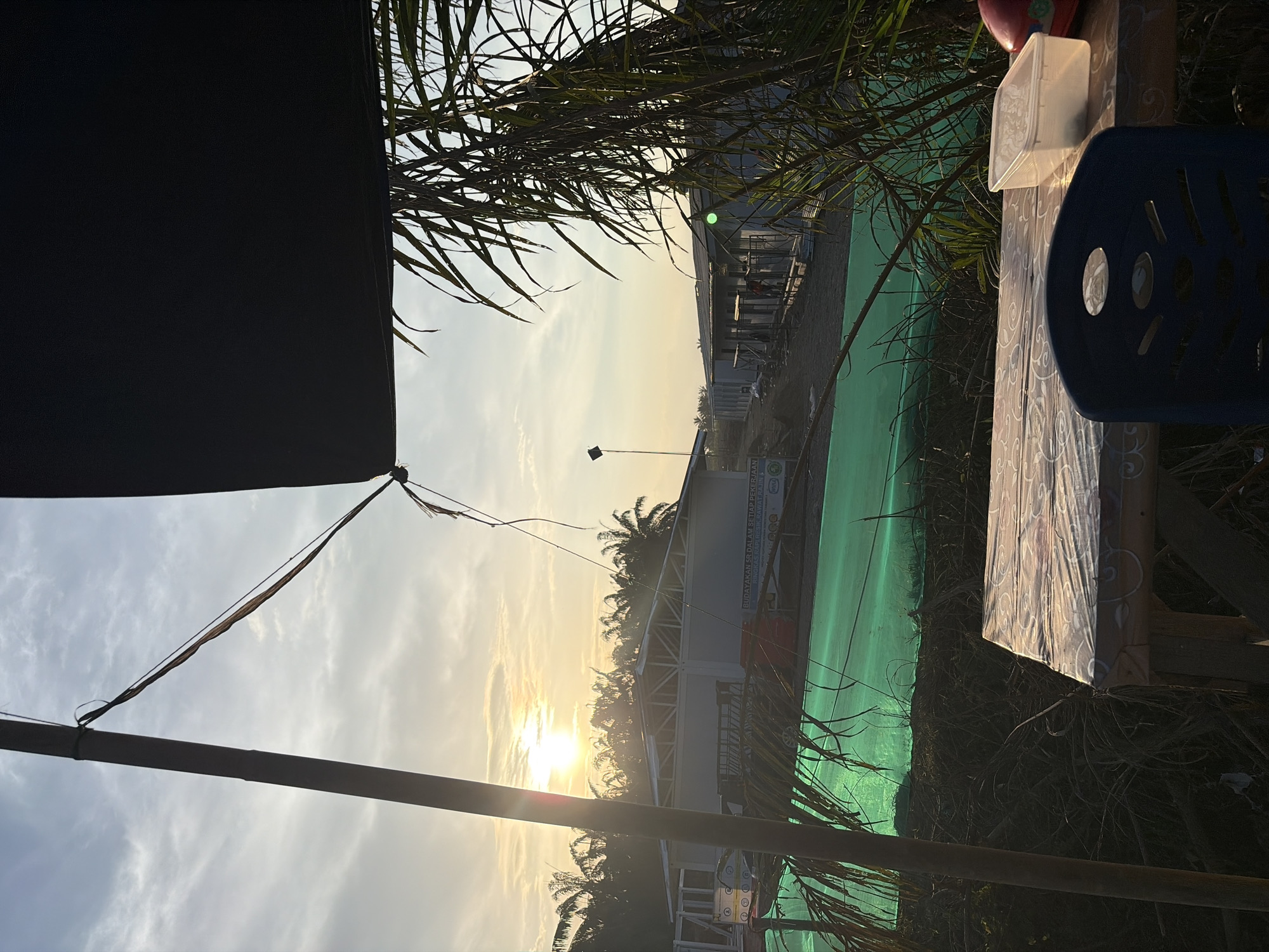 The image depicts a scene in Simpangempat, Indonesia, taken during a sunset. The sun is visible through palm trees and cloudy sky. A temporary structure or tent is partially visible in the foreground, along with a table and chair set. A green tarp or cover is observed in the midground, along with structures that appear to be buildings or modular housing units. There are no people visible.