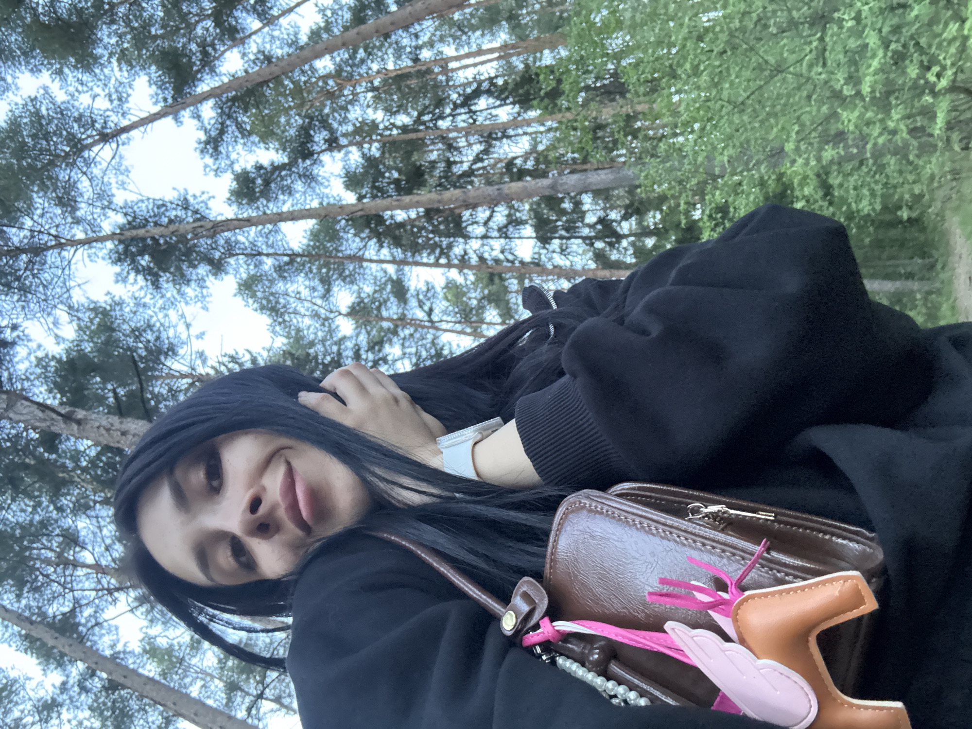 A selfie photograph presents a young woman with black hair, positioned against a backdrop of trees and sky, likely taken outdoors. The woman is wearing a black hooded sweatshirt. A brown purse with pink and tan embellishments is visible. A white watch is on her wrist. The specific location cannot be verified as Votkinsk, Russia.