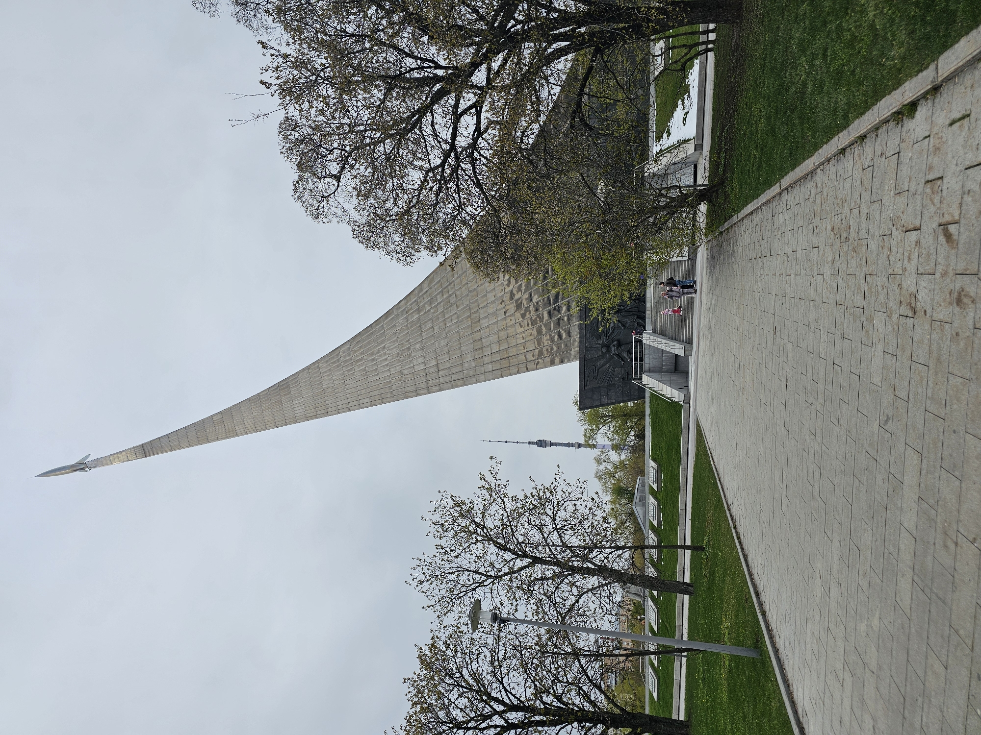 The image captures an outdoor scene featuring the Monument to the Conquerors of Space in Moscow, Russia, under an overcast sky. The monument, a towering, tapering structure, dominates the center-left, culminating in a rocket at its apex. Its surface is textured, appearing metallic or stone-clad. At the base of the monument, a darker, possibly bronze relief sculpture is visible, with a small group of people, including children, standing on steps below it.
To the right, a wide, paved pathway of rectangular light-colored tiles extends from the foreground into the midground, running parallel to the monument. Lush green grass lines the pathway on both sides, with some smaller, subtle structures or plaques embedded in the grass on the left. Several trees with bare branches and emerging light green foliage are scattered throughout the scene, particularly in the foreground and midground, framing the monument. A street lamp post stands near one of these trees in the lower midground.
In the distance, behind the monument and through the trees, the iconic Ostankino Television Tower is distinctly visible against the pale sky. The overall impression is daytime, possibly early spring, given the state of the trees, and the weather is gray and cloudy. No discernible text is clearly visible on any of the structures or signs.
