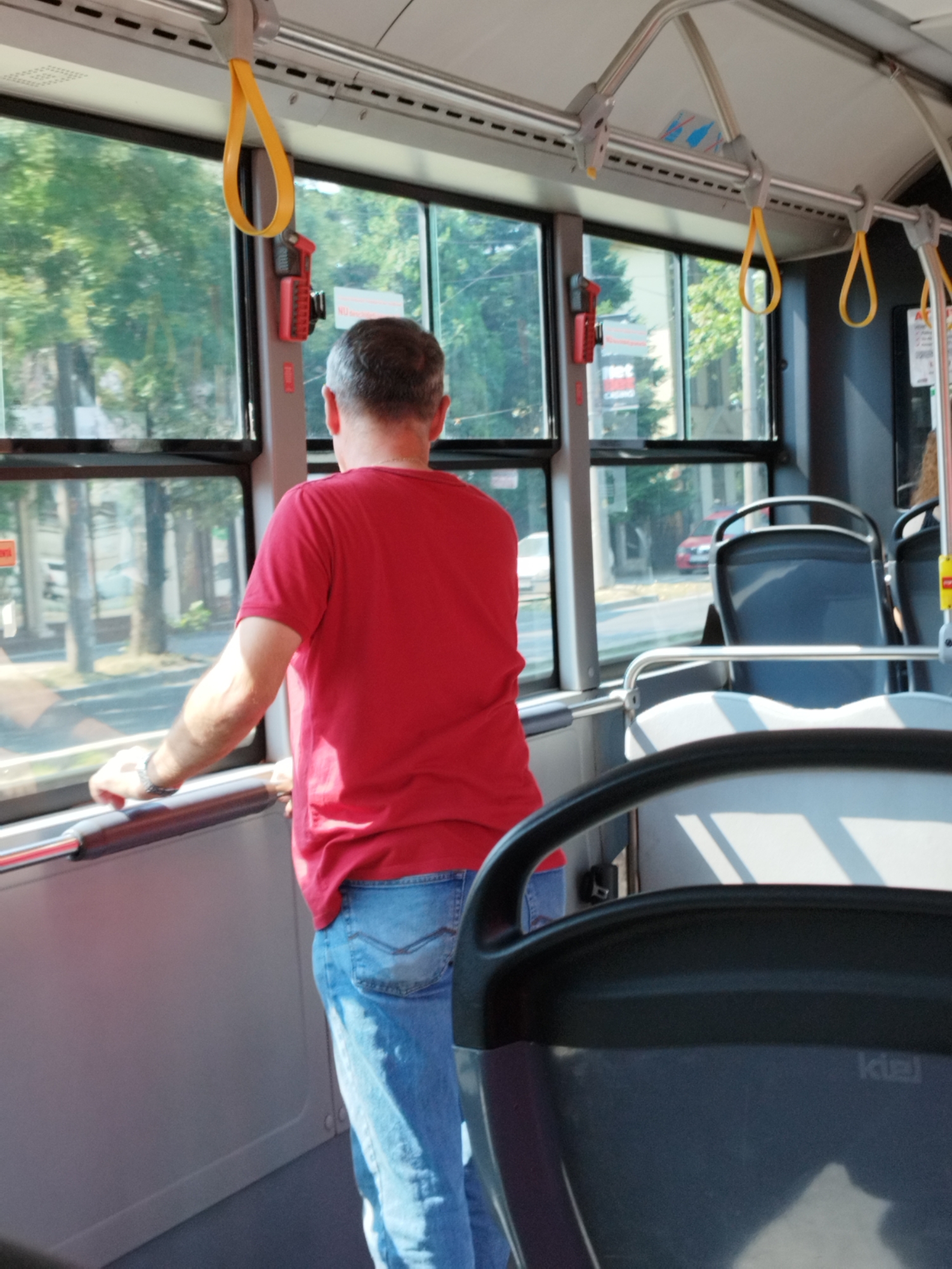 A man in a red t-shirt and blue jeans stands on a bus, looking out the window. He is holding onto a silver pole. The bus is equipped with yellow grab handles and dark-colored seats. Outside the window, trees and buildings are visible. The scene appears to be in Bucharest, Romania, as indicated by the presence of a "Kiel" branded seat, a manufacturer common in public transportation in the region. The lighting suggests it is daytime.