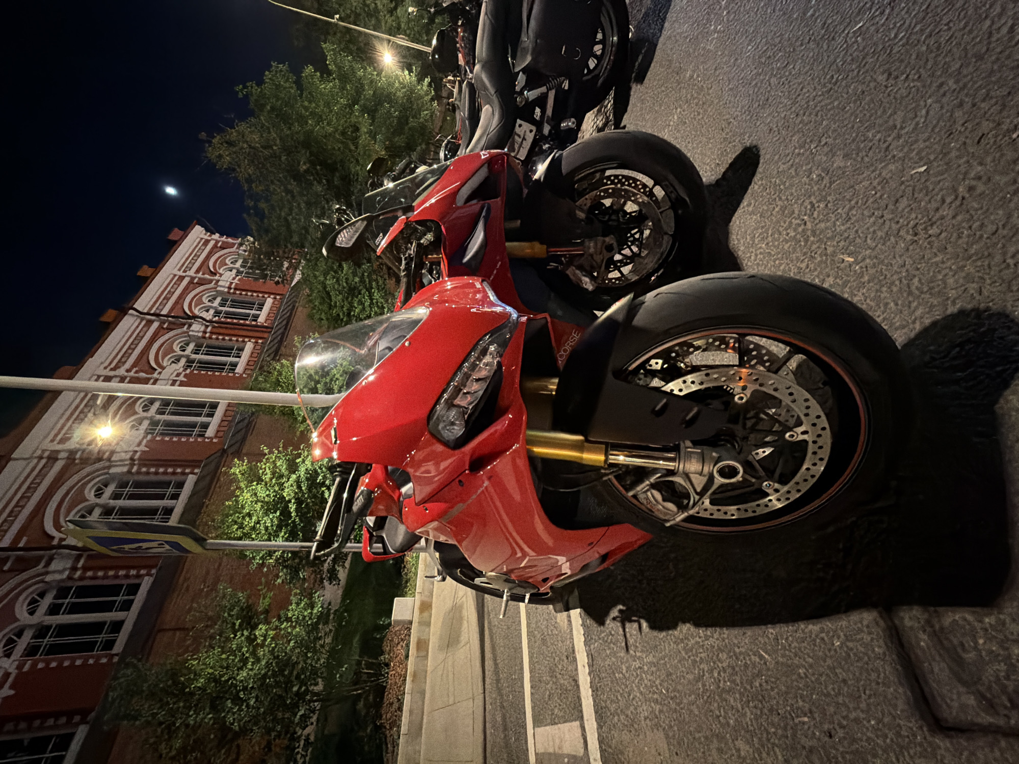 The image shows several parked motorcycles on a street at night in Moscow, Russia. The motorcycles are lined up on the edge of a sidewalk, with the front of a red Ducati visible in the foreground. Behind the Ducati, there are other motorcycles, including a black one. In the background, a building with white arched windows and orange brickwork is visible, illuminated by streetlights. The sky is dark, with a visible moon. A pedestrian crossing sign is visible on the left. The environment is urban, and the scene appears to be captured during a low-light condition.