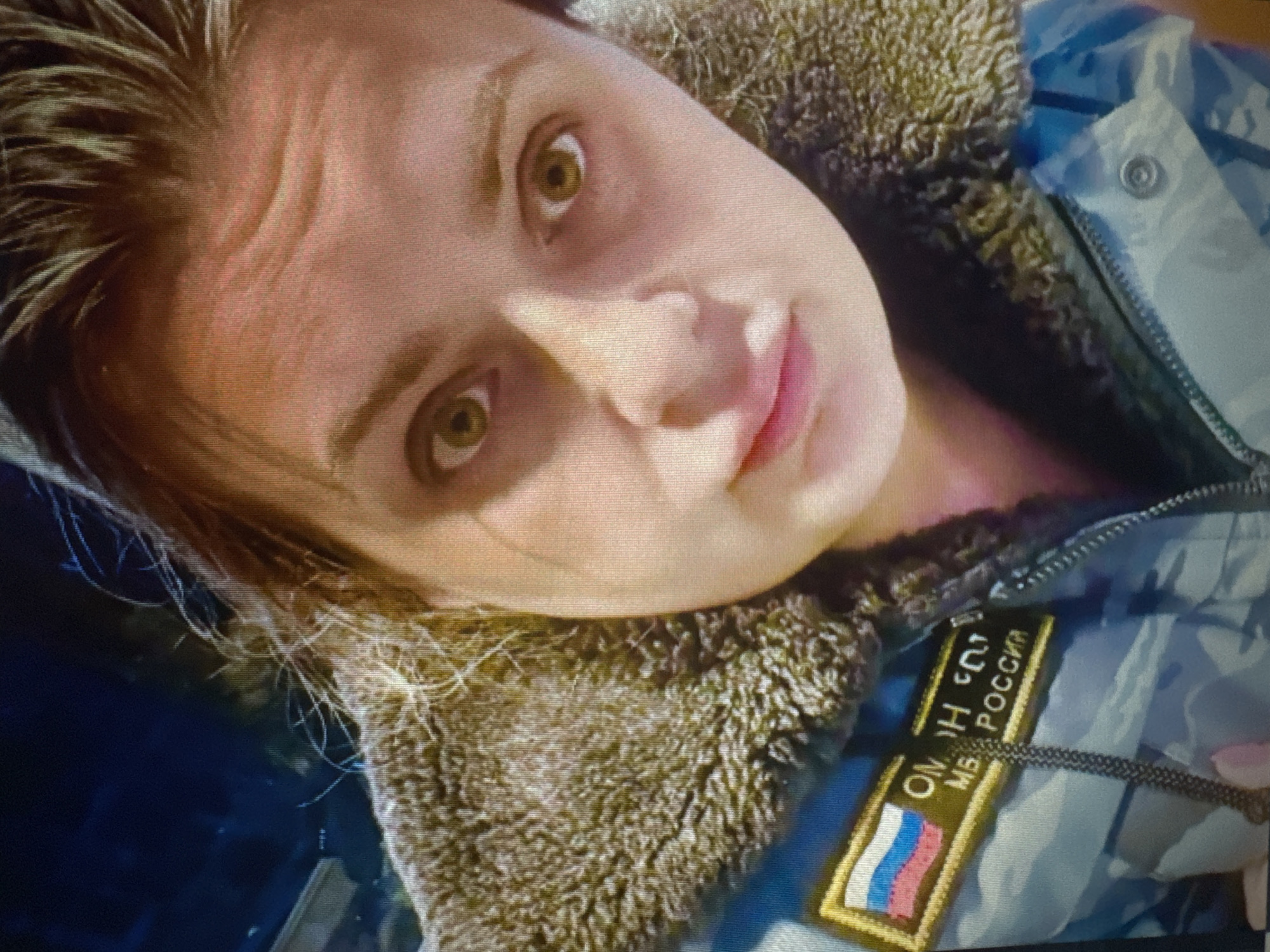 A close-up, waist-up, indoor selfie of a woman with light brown hair and green eyes. She is wearing a camouflage jacket with a fur-lined collar. A patch on the left side of her jacket displays the Russian flag and text that appears to read "ОМОН МВД РОССИИ" which translates to "OMON Ministry of Internal Affairs of Russia". The background is dark, suggesting nighttime or an indoor setting with low lighting. The woman's expression is neutral. The location is specified as Protasovo, Russia.