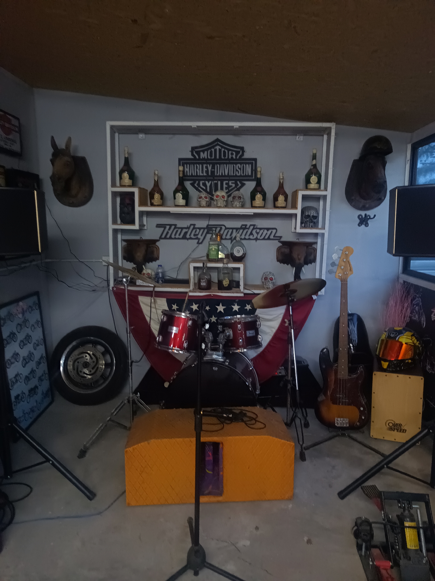 The image shows a cluttered room filled with musical instruments and Harley-Davidson memorabilia. A drum set with red toms sits in front of an American flag draped over a stand. A bass guitar and a cajón drum stand to the right of the drum set. An orange amplifier box sits in the foreground. A motorcycle tire leans against the wall behind the drums. The wall features a white shelf with Harley-Davidson signs, decorative bottles, and skulls. Stuffed animal heads hang on the wall. The location is possibly a music room or garage converted into a practice space, but the precise location in Surat Thani City Municipality, Thailand, cannot be determined from the image. There are no people visible in the image.