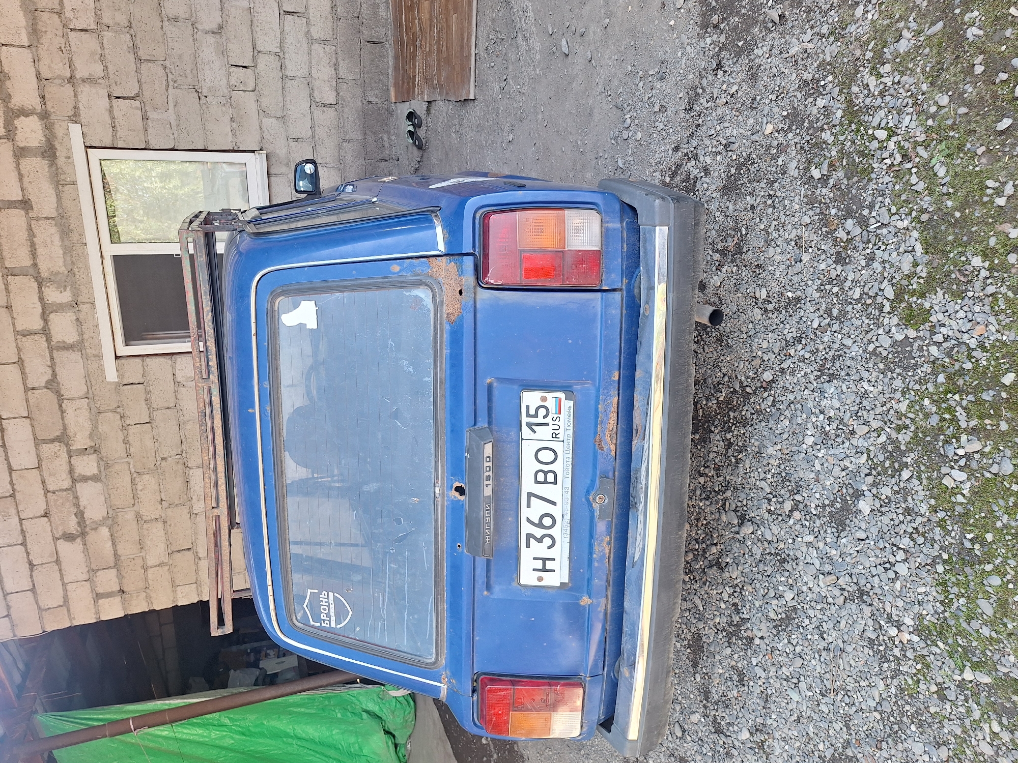The image depicts the rear of a blue, older model car parked on a gravel surface next to a brick building in Chermen, Russia. The car has a roof rack and visible rust spots. The building has a window and a green tarp hanging on its left side. A license plate with the characters "H 367 BO 15" is attached to the rear of the car. The ground is covered in a mix of gravel and sparse grass. There are no people visible in the image.