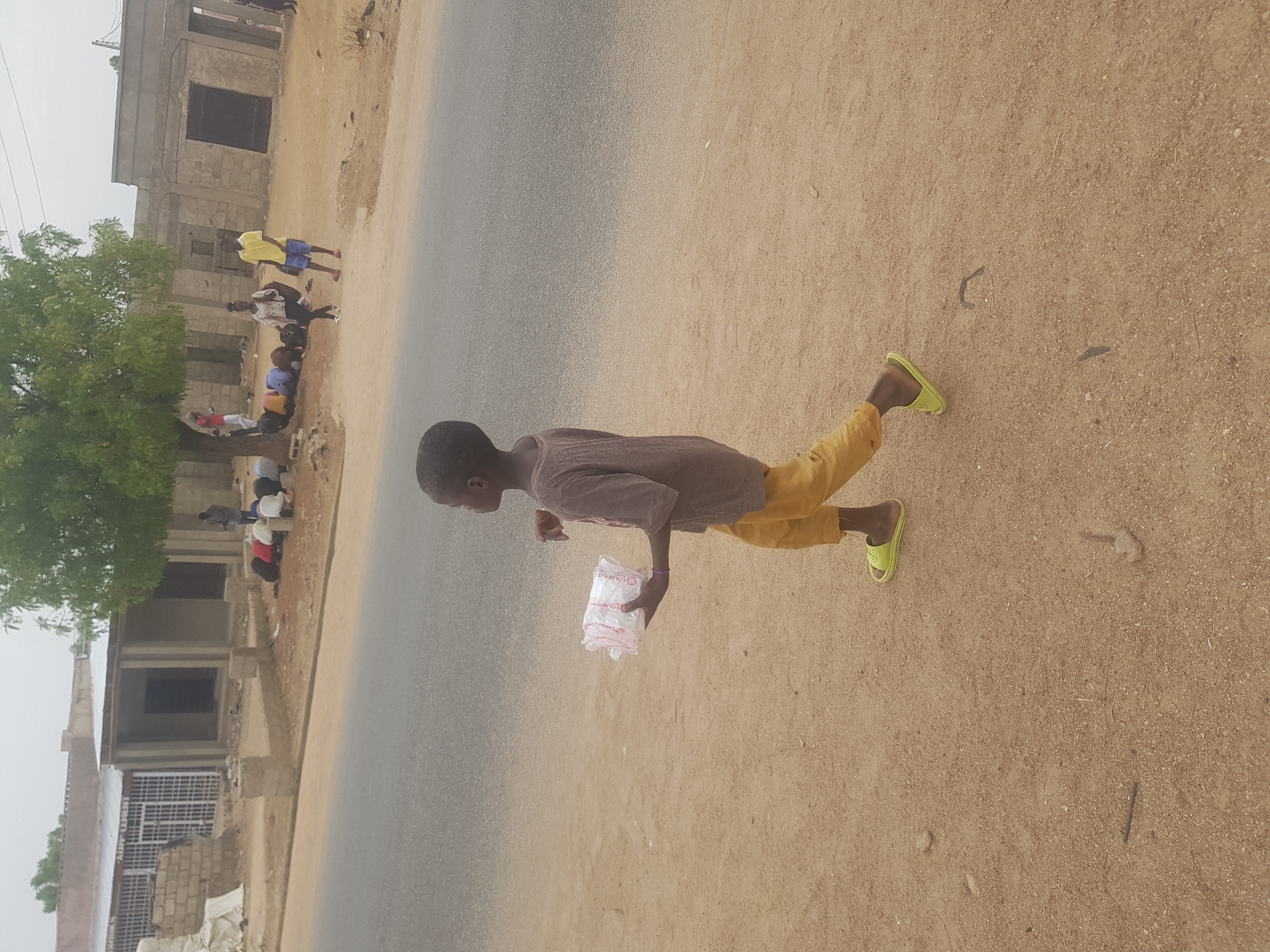 A young person is walking on a dirt shoulder next to a paved road in Dutsinma B, Nigeria. The person is wearing a gray shirt, yellow pants, and green sandals, and holding a plastic bag. On the left side of the road are a partially constructed building and a group of people gathered under a tree.