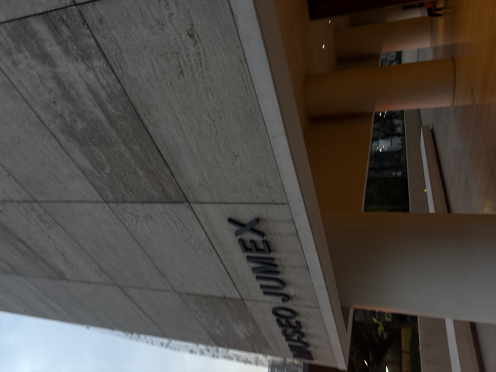 The image captures an upward angle view of the Museo Jumex, located in Mexico City, Mexico. The focus is on the museum's facade, constructed from light-gray, horizontally-laid panels, with the museum's name, "MUSEO JUMEX," spelled out in block letters. To the right of the building's facade, smooth, cylindrical columns support the structure, giving glimpses into the interior spaces and views of the outside. The photograph appears to be taken during dusk or dawn, with softer lighting and shadows accentuating the building's architectural details.