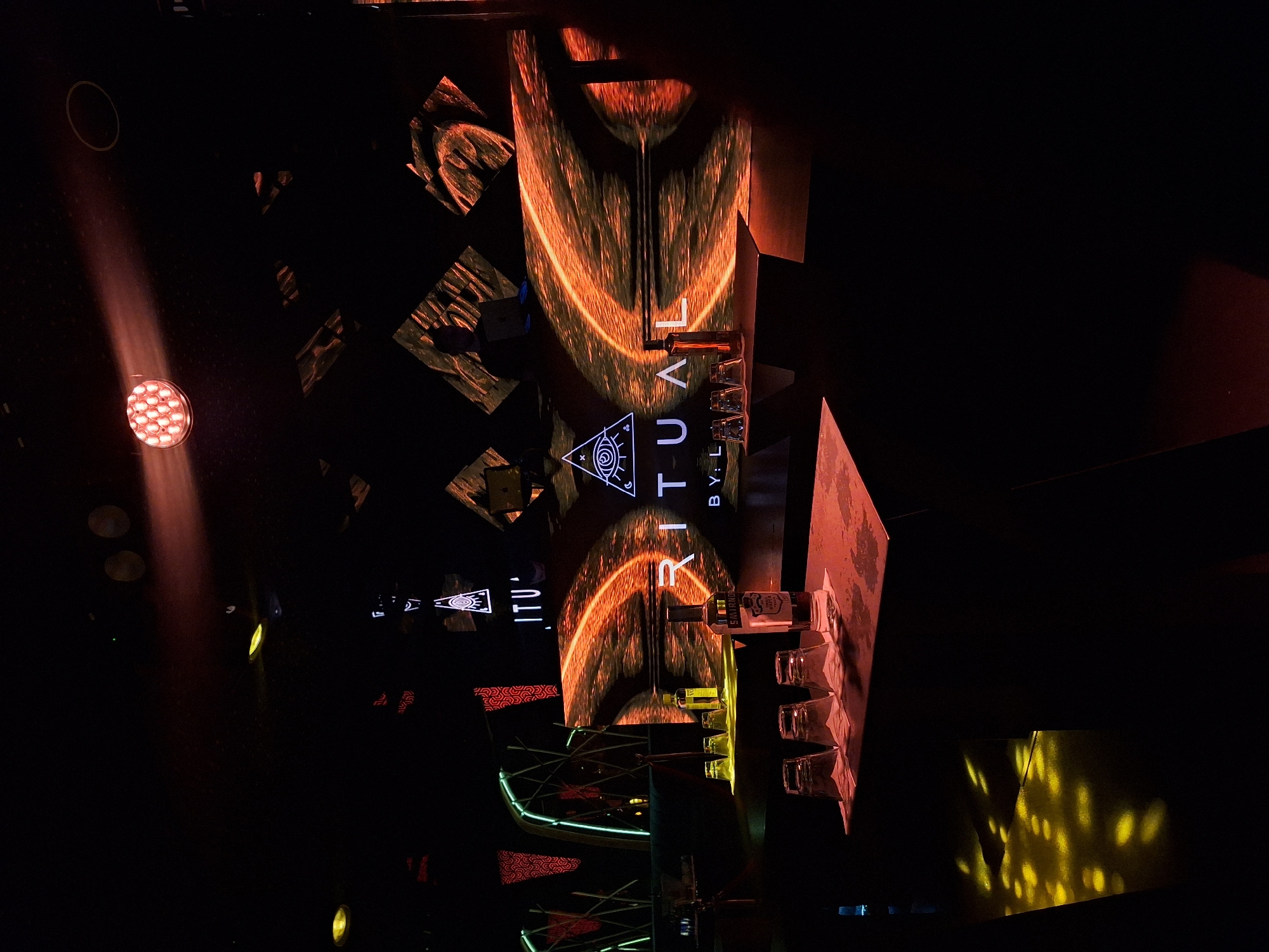 The image, oriented correctly, depicts an indoor venue at night, likely a bar, club, or event space in Sofia, Bulgaria, characterized by dynamic lighting and projections. The scene is dark, with strong artificial illumination creating a vibrant, energetic atmosphere. No people are visible.
The central focus is a large, vertical display or screen projecting the word "RITUAL" in white, stylized letters, centered around a symbol resembling an Eye of Providence. Below "RITUAL," the text "BY: L" is also visible. The background of this projection features warm-toned, abstract, flowing patterns of orange and black, possibly resembling molten liquid or cosmic imagery.
In front of this main display, on a surface below, several clear glasses and at least two bottles are arranged. One bottle prominently displays a label with the brand name "SANTANA TRIPLE DISTILLED."
The surrounding environment includes various lighting fixtures and architectural elements. To the left, a bright red spotlight with multiple LED bulbs casts a warm beam across the dark space. Other yellow circular lights are visible further back. Geometric structures, some illuminated with green neon-like lighting, are arranged throughout the space, adding to the modern, abstract aesthetic. Triangular patterns with red and white designs are also seen on vertical surfaces. On the right side, dappled yellow light patterns are projected onto a dark wall or floor, creating a textured visual effect. The overall impression is that of a contemporary and stylish venue designed for social gatherings or entertainment.