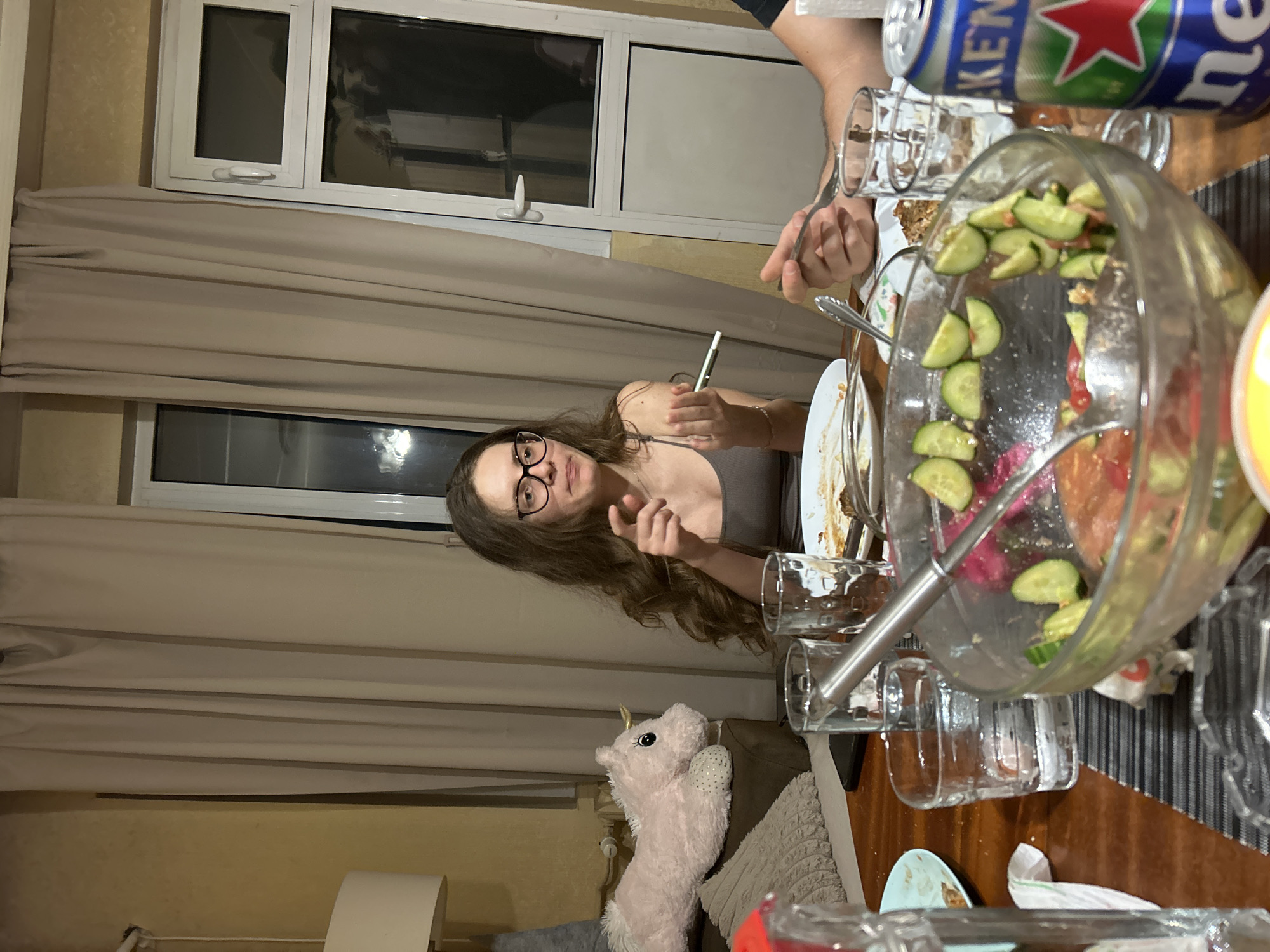 A woman with long, wavy brown hair and glasses is seated at a table in a dining room, gesturing with her left hand. She is wearing a grey tank top. The table is set for a meal, with plates, glasses of water, and a salad bowl containing sliced cucumbers and tomatoes. A can of Heineken is visible on the right side of the table. In the background, beige curtains cover a window, and a pink unicorn stuffed animal is on a couch. The scene appears to be a casual gathering or meal. The location is not specified to be in Sofia, Bulgaria, but it is an indoor residential setting.